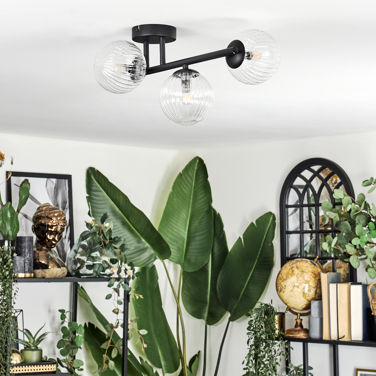 Ein modernes Zimmer hat eine Deckenlampe mit Glaskugeln. Große grüne Pflanzen, Regale mit Dekoration und ein Globus sind im Raum.