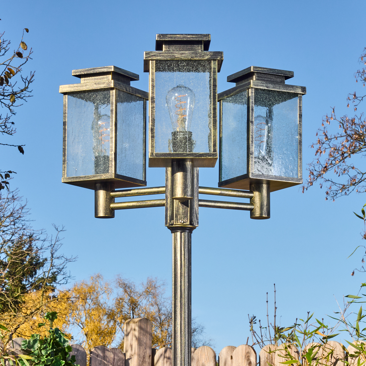 Eine dekorative Außenlampe mit drei Lichtern. Der Himmel ist blau. Im Hintergrund sind ein Holzzaun und einige Bäume.