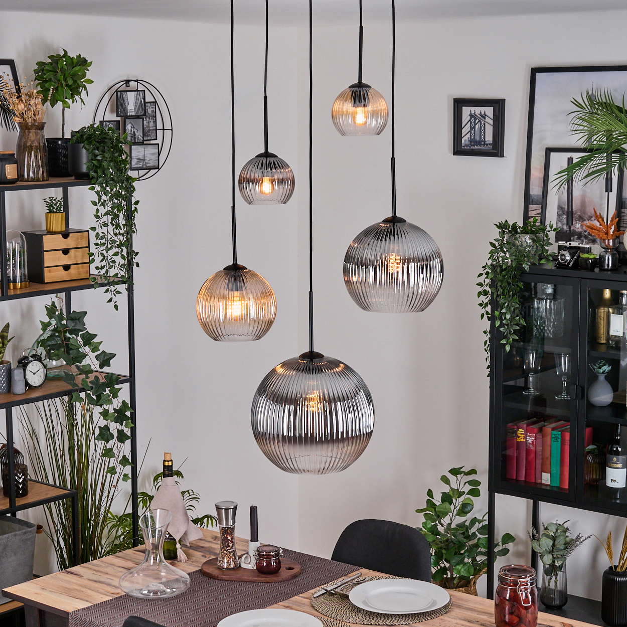 Esszimmer mit Holztisch, Hängelampen, schwarzen Regalen, Pflanzen und einem schwarzen Schrank mit verschiedenen Gegenständen.