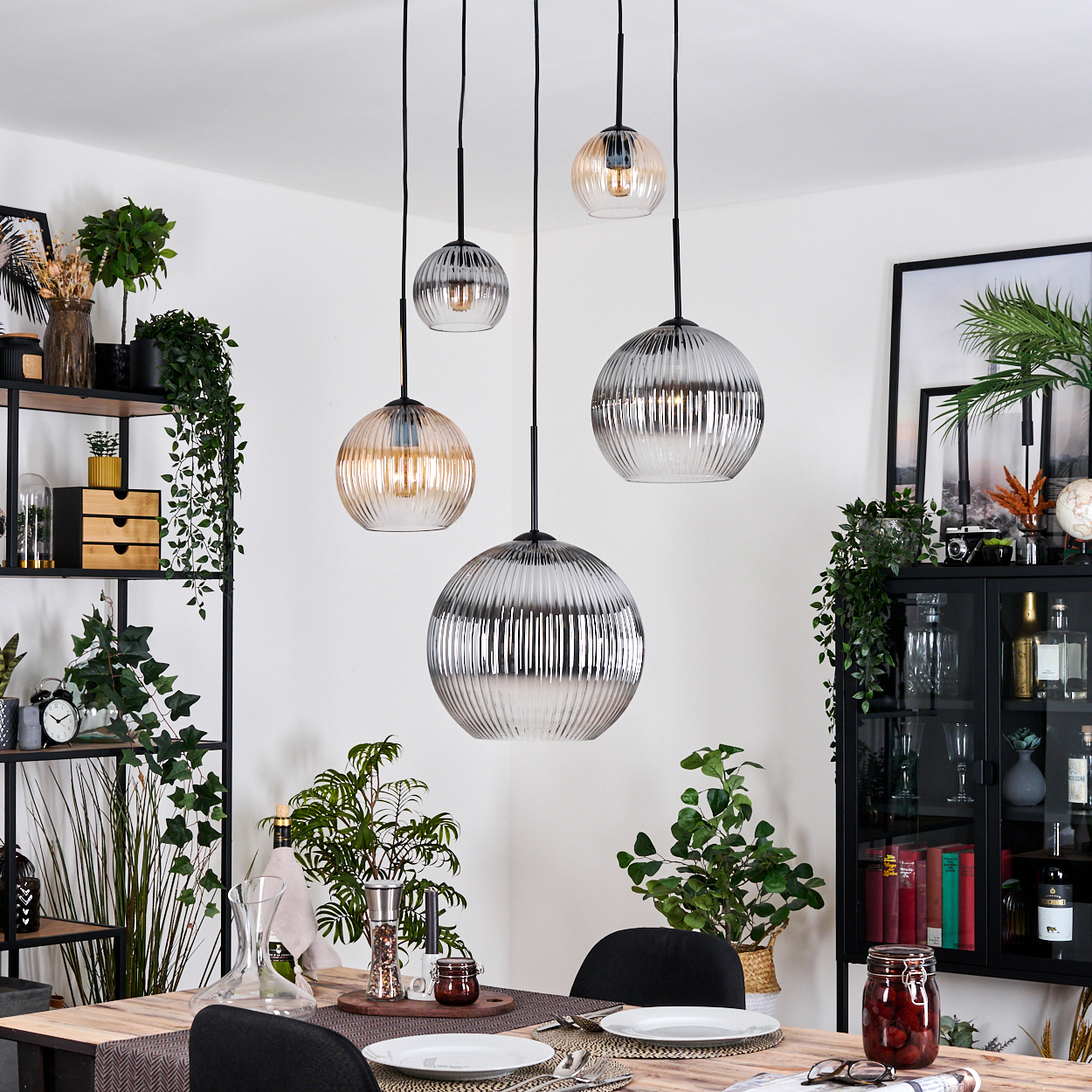 Esszimmer mit hängenden Glaslampen. Tisch mit Tellern gedeckt. Regale mit Pflanzen und Dekor. Schwarzer Schrank mit Flaschen.