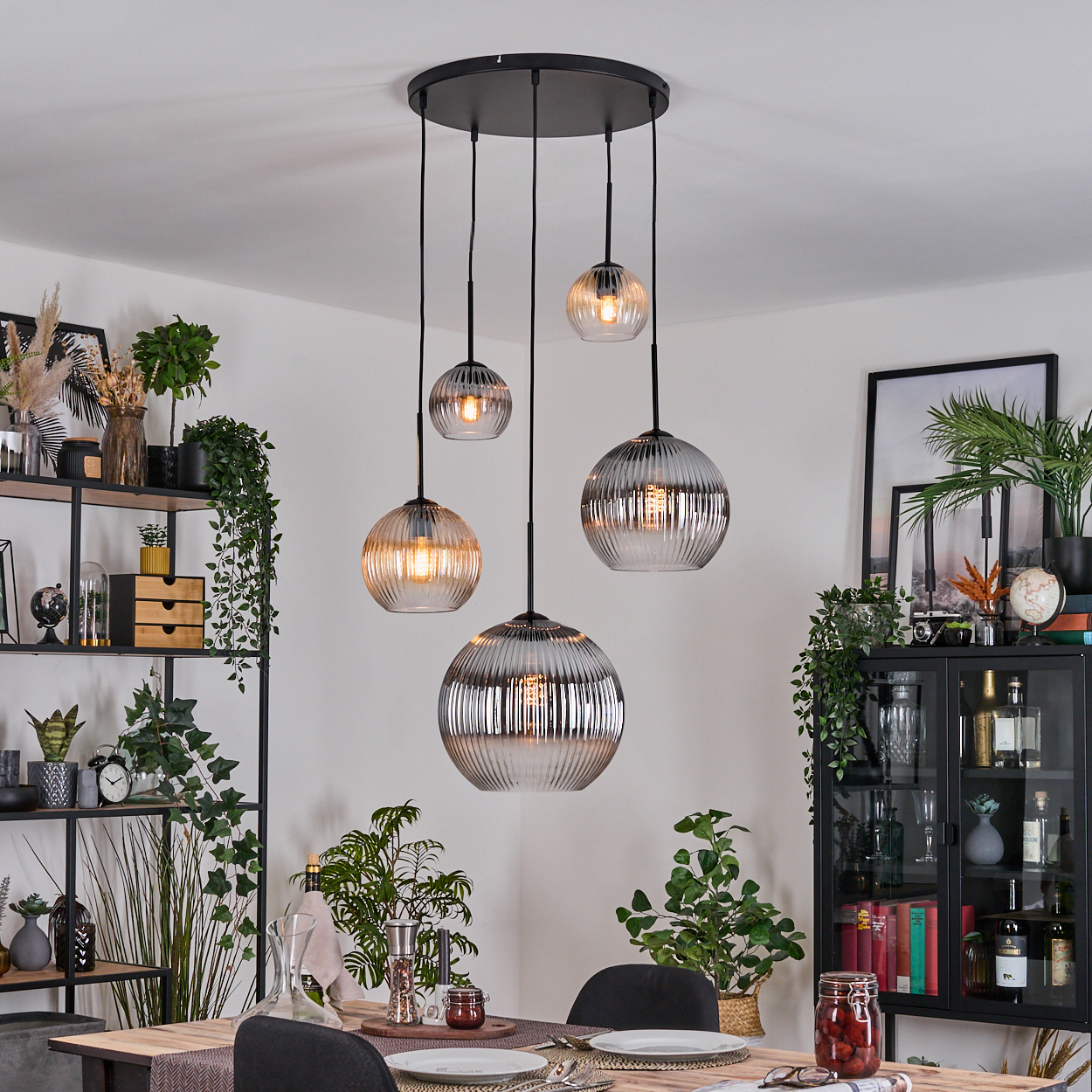 Esszimmer mit hängenden Kugelleuchten. Tisch mit Tellern gedeckt. Regale und Schrank mit Pflanzen und Dekor. Pflanzen in Töpfen.