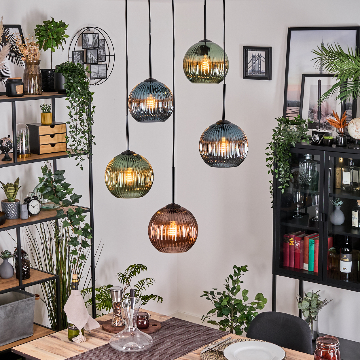 Esszimmer mit hängenden Kugelleuchten. Tisch mit Tellern, Pflanzen und Regalen mit Dekorationen. Schwarzer Schrank mit Büchern.