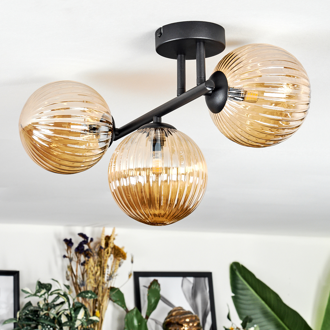 Eine moderne Deckenlampe mit drei bernsteinfarbenen Glaskugeln. Schwarzer Metallrahmen. Im Hintergrund sind Pflanzen und gerahmte Bilder zu sehen.