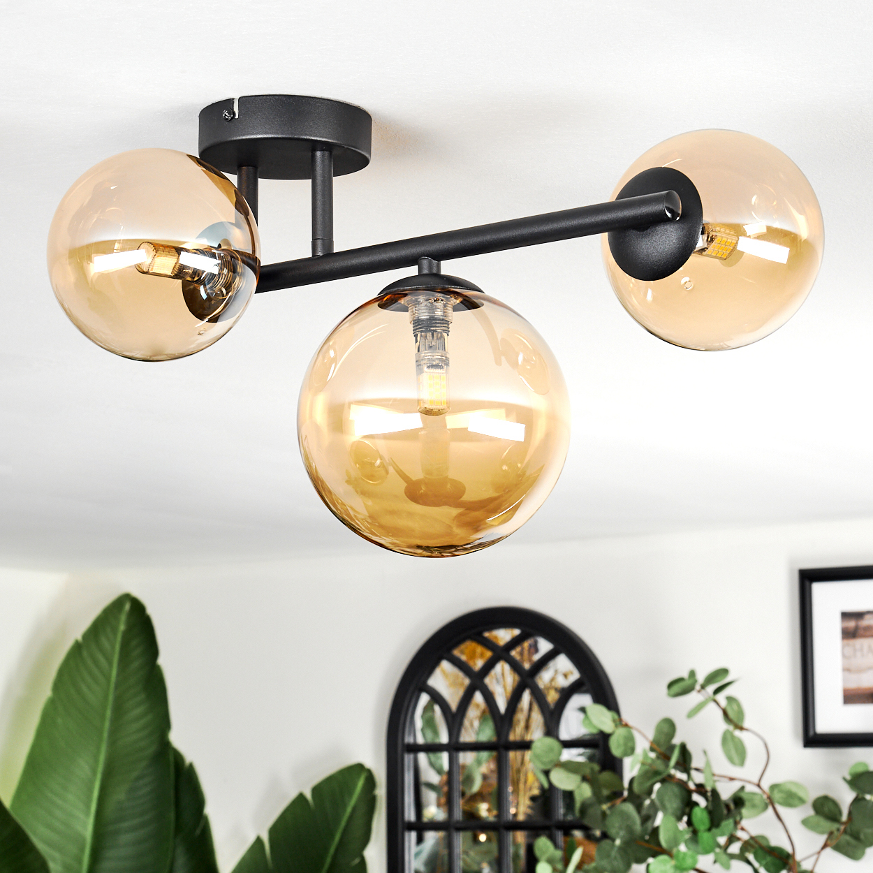 Eine moderne Deckenlampe mit drei bernsteinfarbenen Glaskugeln. Schwarzer Metallrahmen. Pflanzen und ein dekoratives Fenster sind im Hintergrund zu sehen.