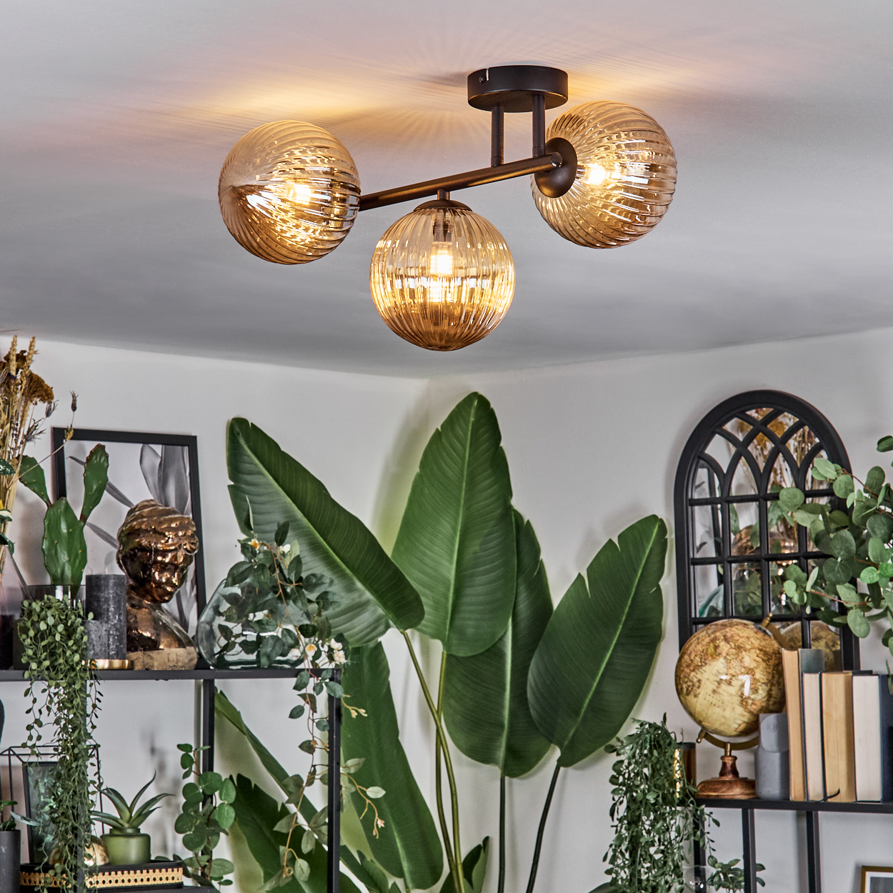 Deckenlampe mit drei Kugelschirmen, grüne Pflanzen, Regale mit Dekoration und ein Globus. Der Raum hat eine weiße Wand.