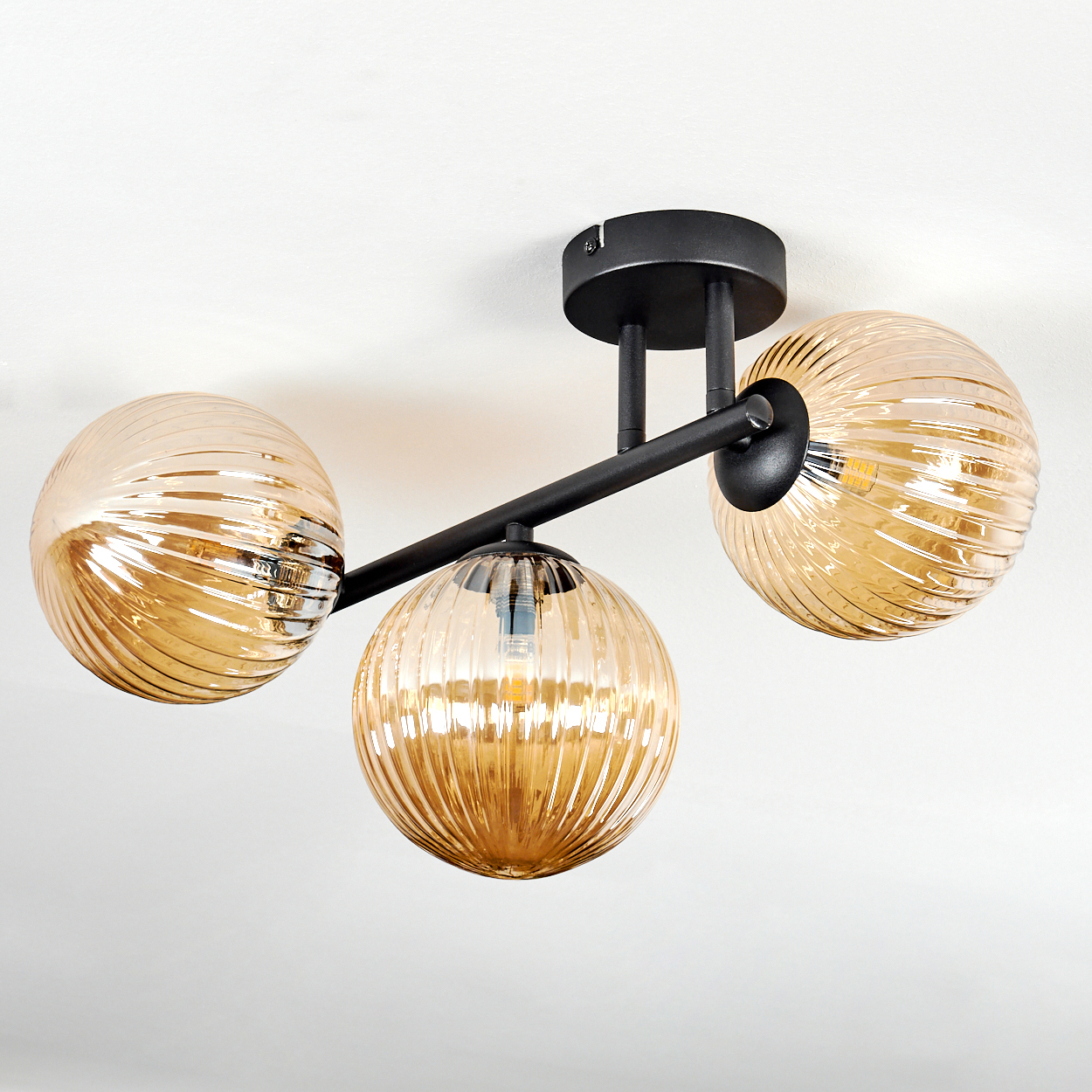 Moderne Deckenlampe mit drei bernsteinfarbenen, kugelförmigen Glasschirmen. Schwarze Metallarme und Deckenhalterung. Weißer Deckenhintergrund.