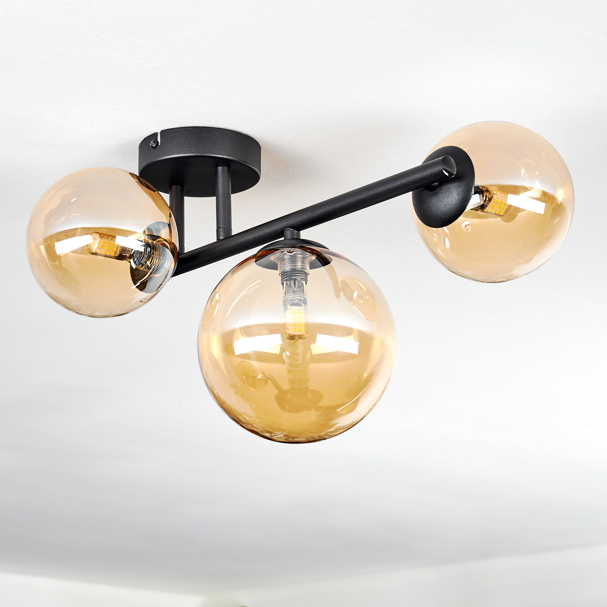 Eine moderne Deckenleuchte mit drei bernsteinfarbenen Glaskugeln. Sie hat einen schwarzen Metallrahmen und ist an einer weißen Decke befestigt.