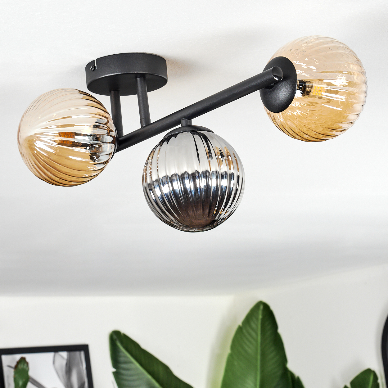 Moderne Deckenleuchte mit drei kugelförmigen Schirmen. Zwei sind bernsteinfarben, einer verspiegelt. Schwarze Metallarme und -basis. Weißer Deckenhintergrund.