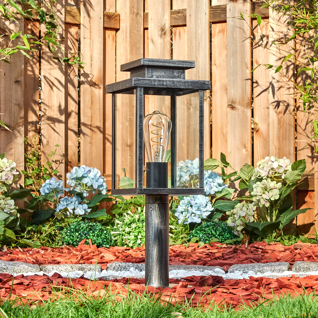 Eine dekorative Außenlampe mit einer Vintage-Glühbirne steht in einem Garten. Der Hintergrund zeigt einen Holzzaun und verschiedene Pflanzen.