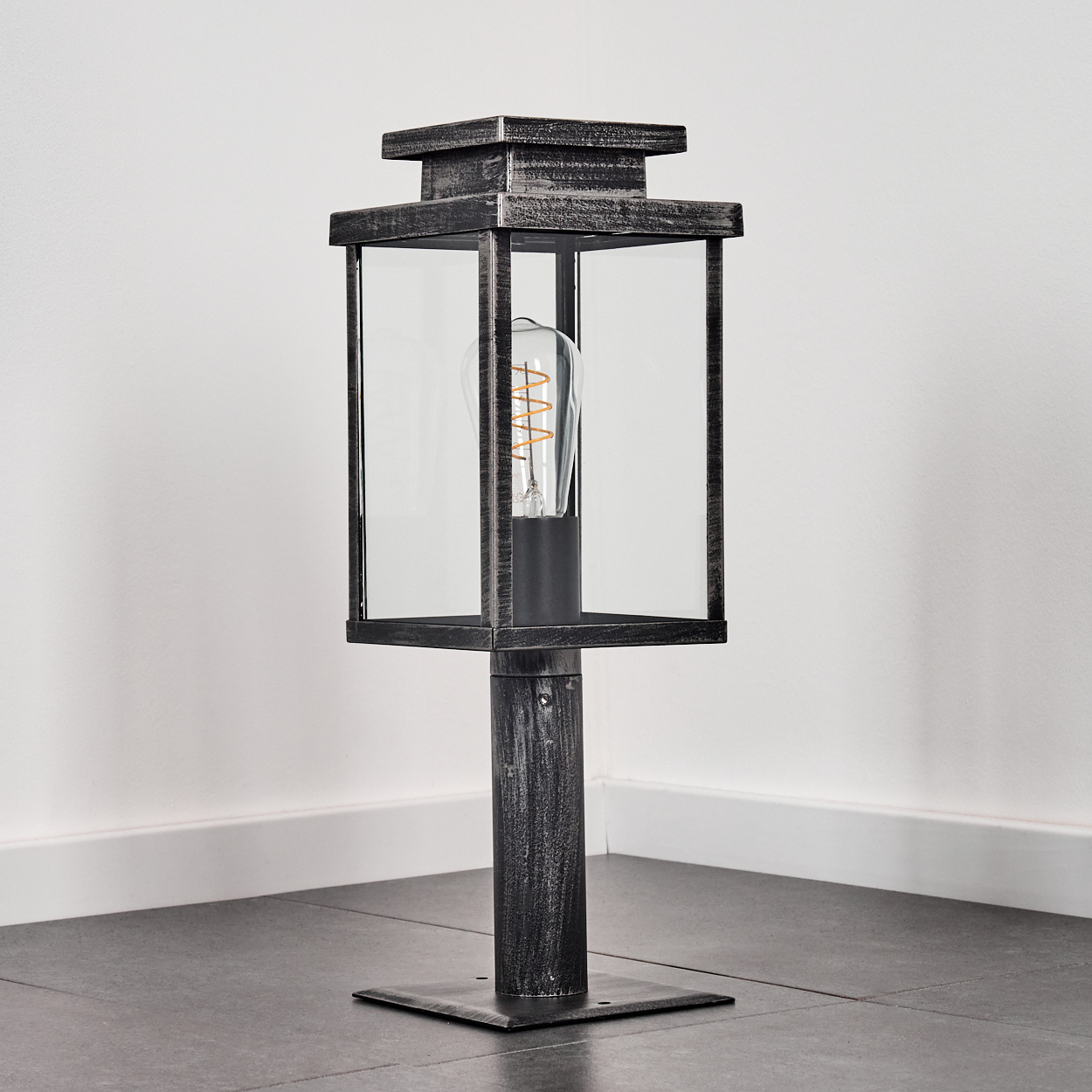 Eine Metall-Außenlampe mit Glasgehäuse und Glühbirne. Sie steht auf einem dunklen Fliesenboden, in der Nähe einer weißen Wand.