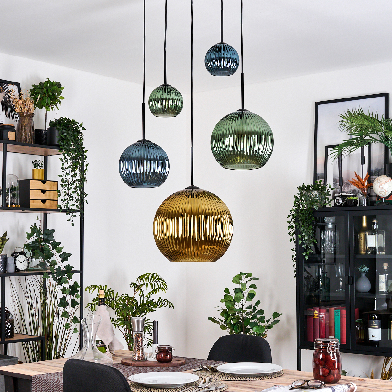 Esszimmer mit hängenden Glaslampen. Tisch für das Abendessen gedeckt. Regale mit Pflanzen und Dekorationsartikeln. Schwarzer Schrank mit Glaswaren.