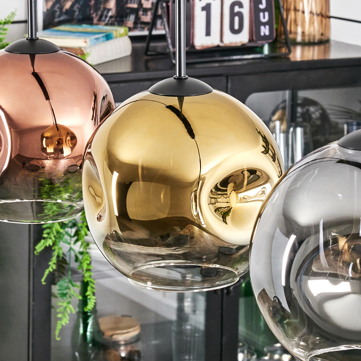 Drei kugelförmige Pendelleuchten in Kupfer, Gold und Silber. Im Hintergrund sind ein Kalender, Pflanzen und Möbel zu sehen.