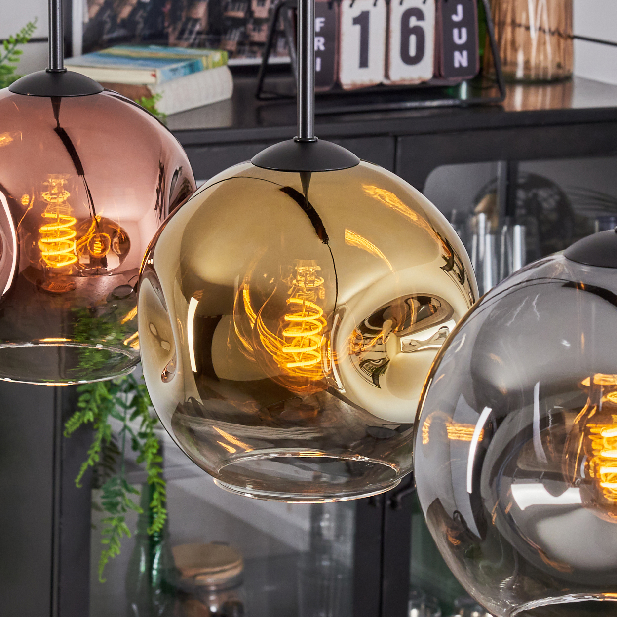 Drei kugelförmige Pendelleuchten in Kupfer, Gold und Grau. Jede mit einer leuchtenden Wendelglühbirne. Der Hintergrund zeigt Dekoration und einen Kalender.