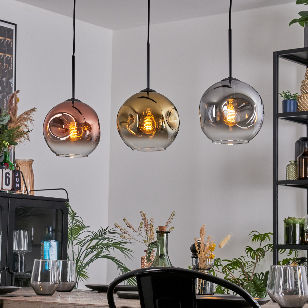 Drei Pendelleuchten mit Glaskugeln, Kupfer, Gold und Grau. Ein Esstisch ist mit Gläsern und einem schwarzen Stuhl gedeckt.