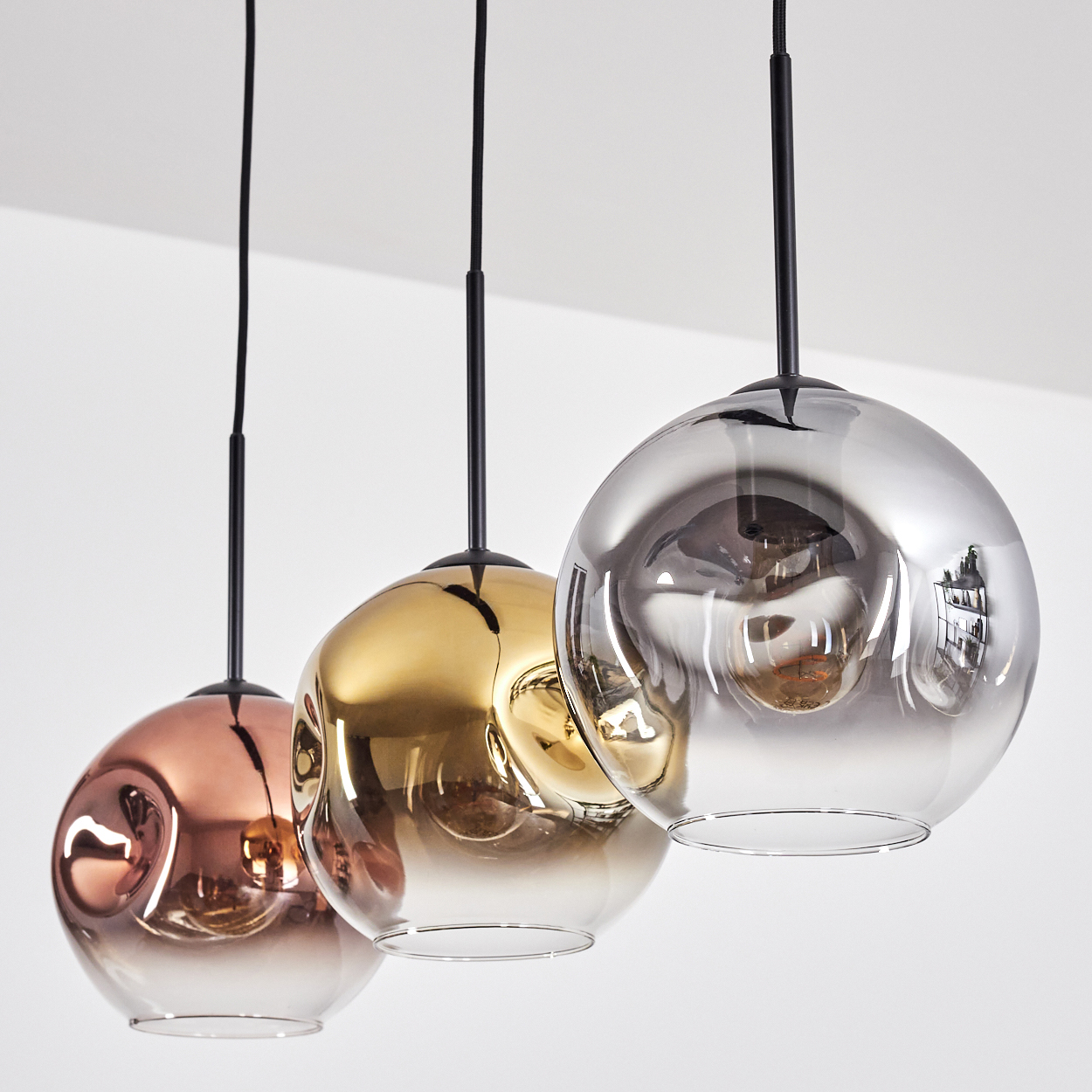 Drei kugelförmige Pendelleuchten in Kupfer, Gold und Silber. Sie hängen an schwarzen Kabeln vor weißem Hintergrund.