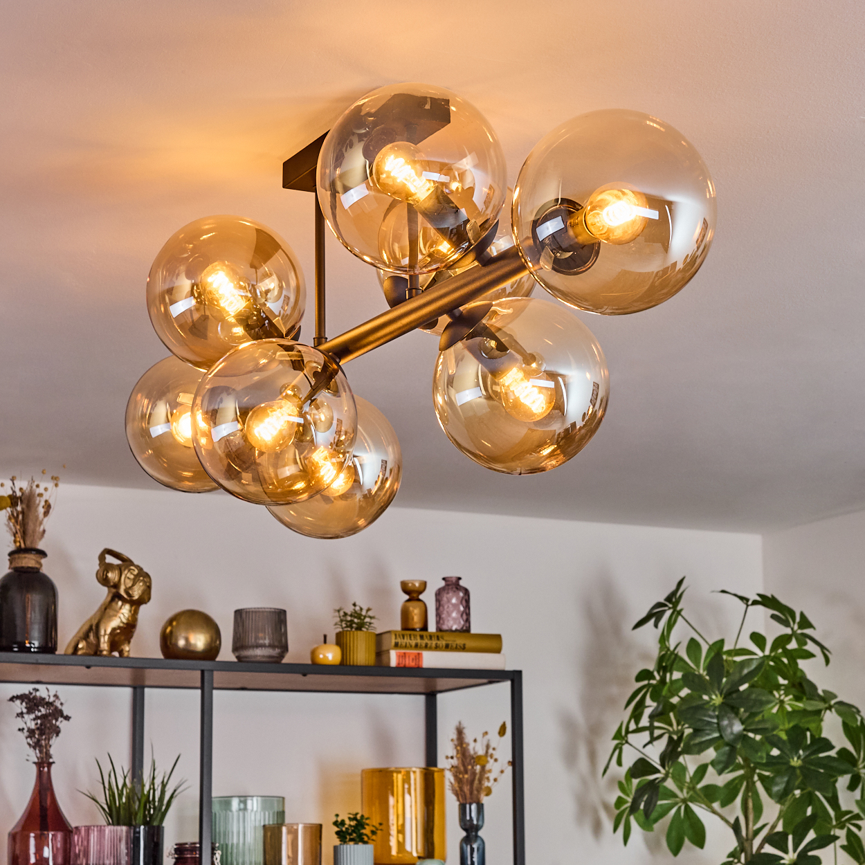 Moderne Deckenleuchte mit bernsteinfarbenen Glaskugeln. Darunter ein Regal mit Dekorationsartikeln und Pflanzen.
