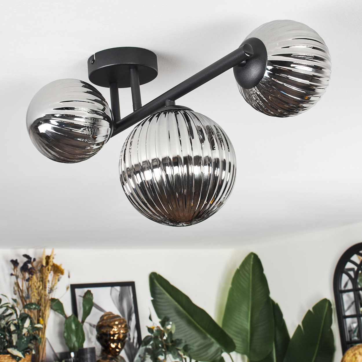 Moderne Deckenlampe mit drei kugelförmigen Schirmen. Die Lampe ist silberfarben, die Decke weiß. Pflanzen und ein Bilderrahmen sind sichtbar.