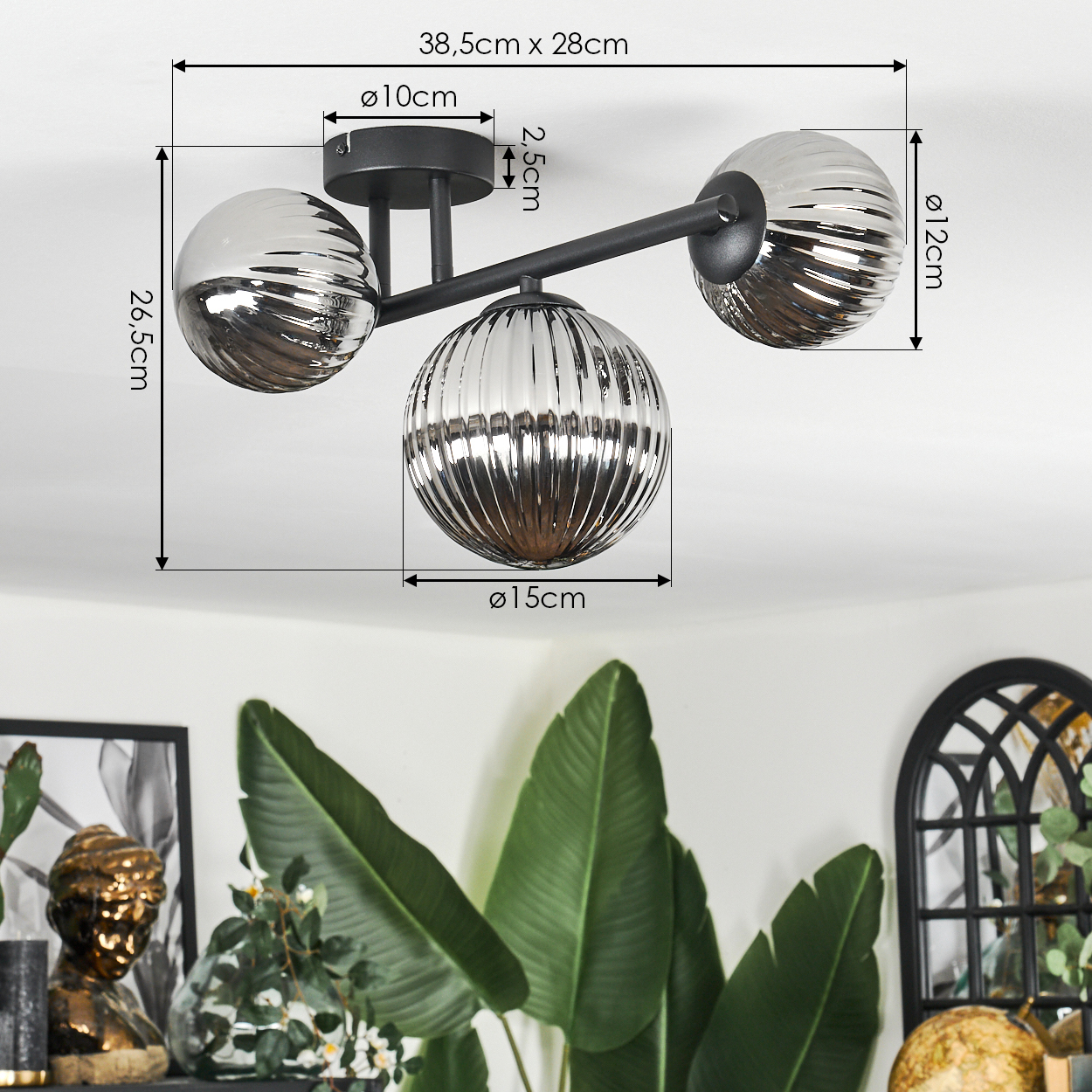 Moderne Deckenlampe mit drei unterschiedlich großen Glaskugeln, schwarzen Armen und Abmessungen. Grüne Pflanzen im Vordergrund.