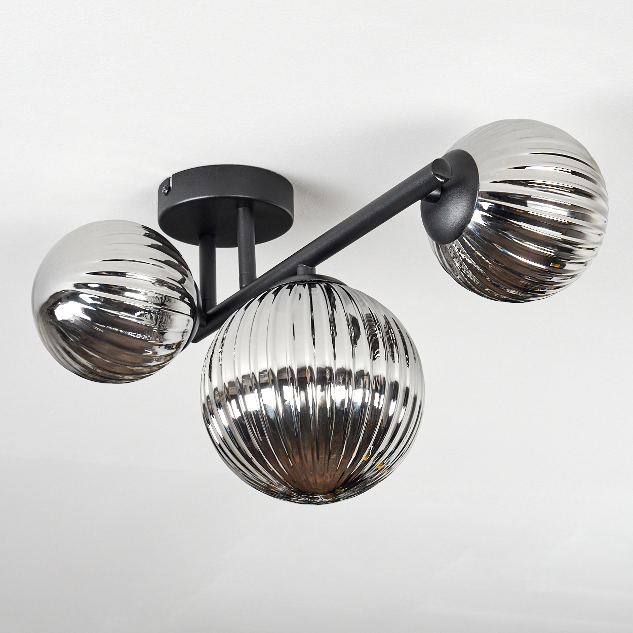 Eine moderne Deckenleuchte mit drei silbernen, gerippten Glaskugelschirmen und schwarzen Metallarmen. Der Hintergrund ist eine weiße Decke.