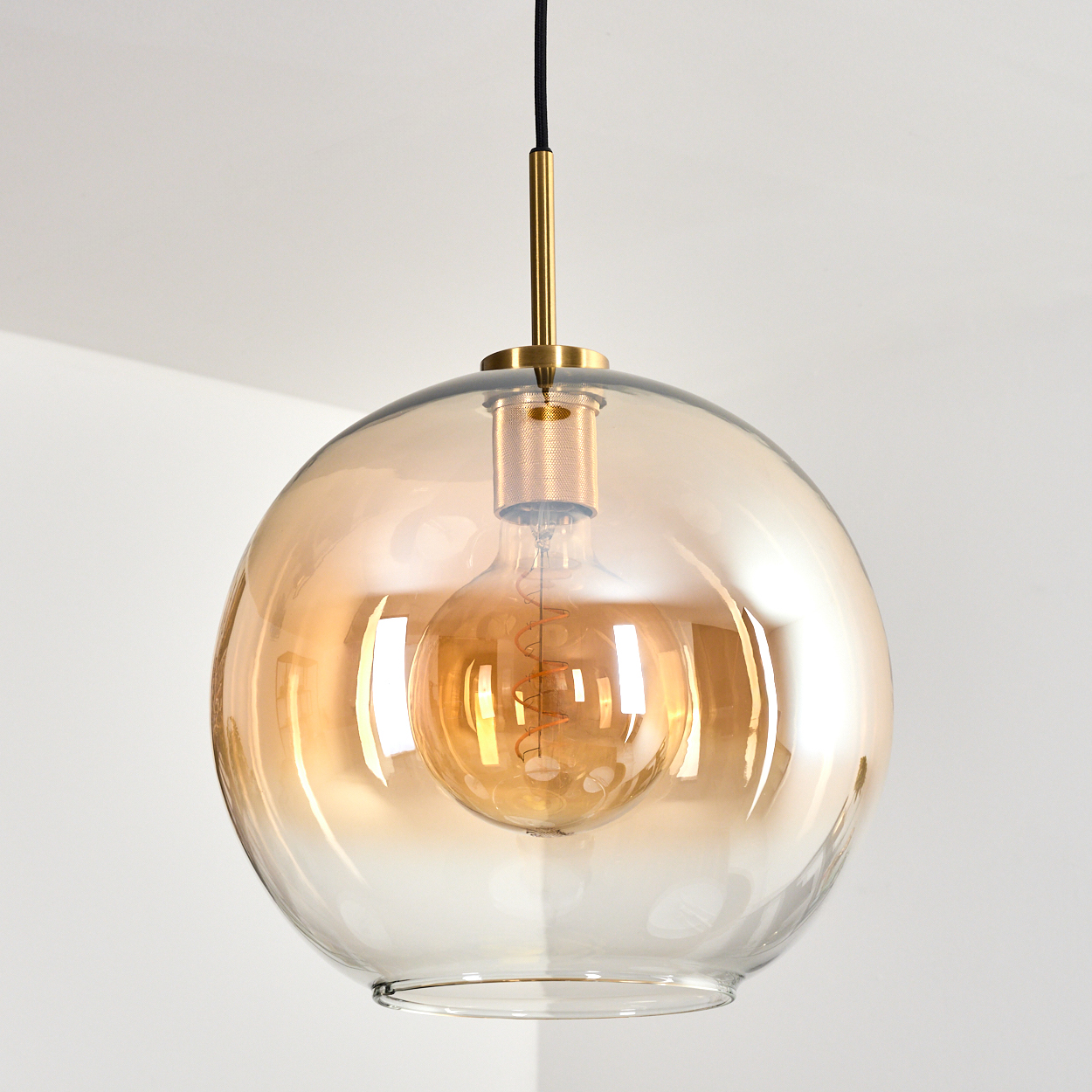 Eine moderne Pendelleuchte mit bernsteinfarbenem Glasschirm. Im Inneren ist eine warm getönte Glühbirne zu sehen. Die Leuchte hängt an einer weißen Decke.