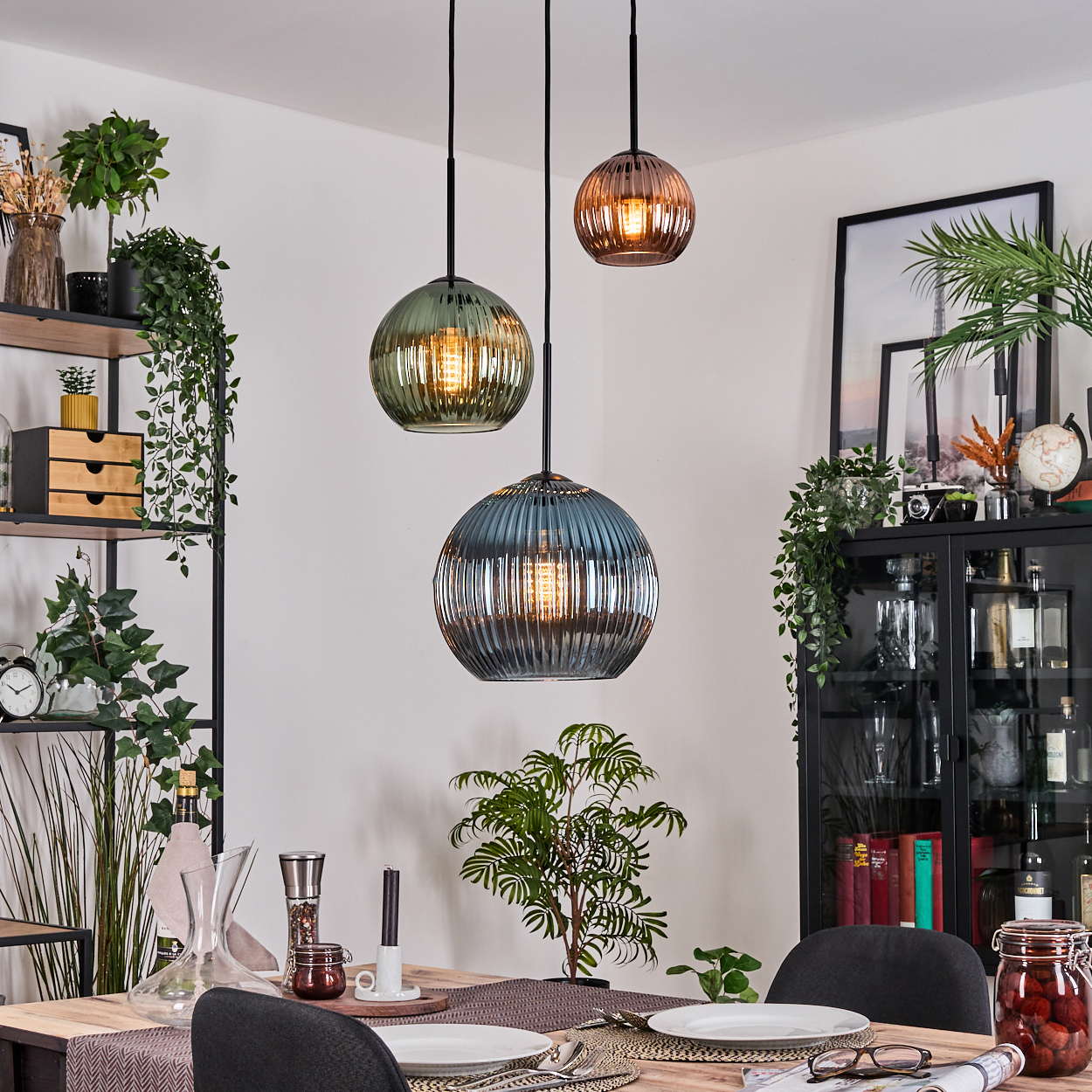 Esszimmer mit drei Pendelleuchten. Tisch mit Tellern gedeckt. Regale, Pflanzen und ein Schrank im Hintergrund.