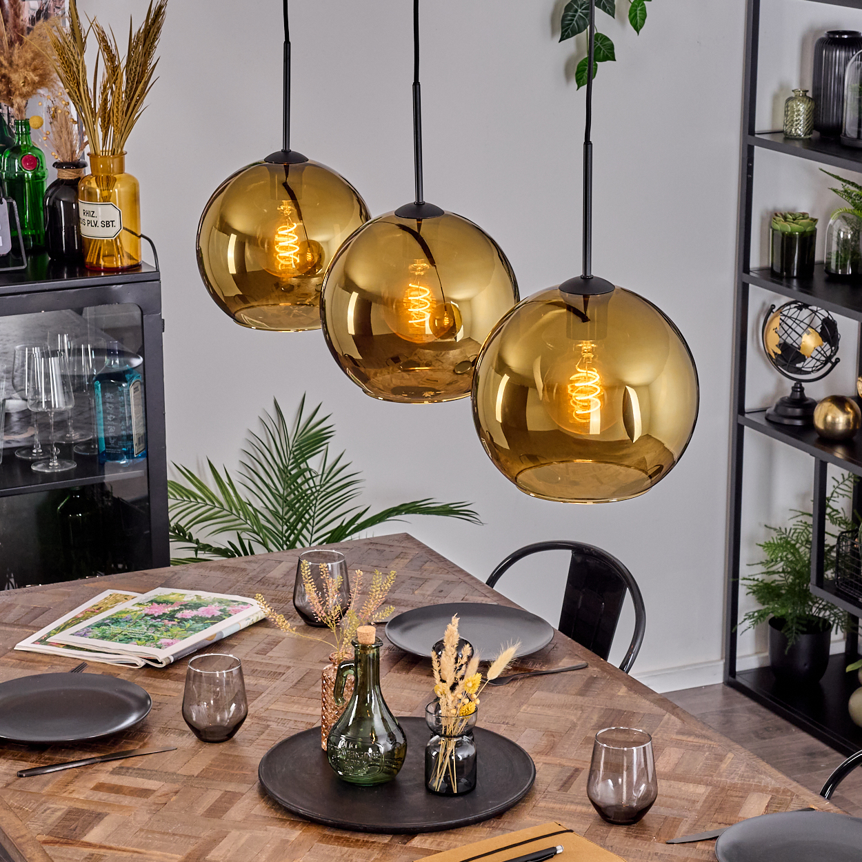 Drei goldene Pendelleuchten über einem Holztisch, der für das Abendessen gedeckt ist. Geschirr, Gläser und Dekorationsartikel sind vorhanden.