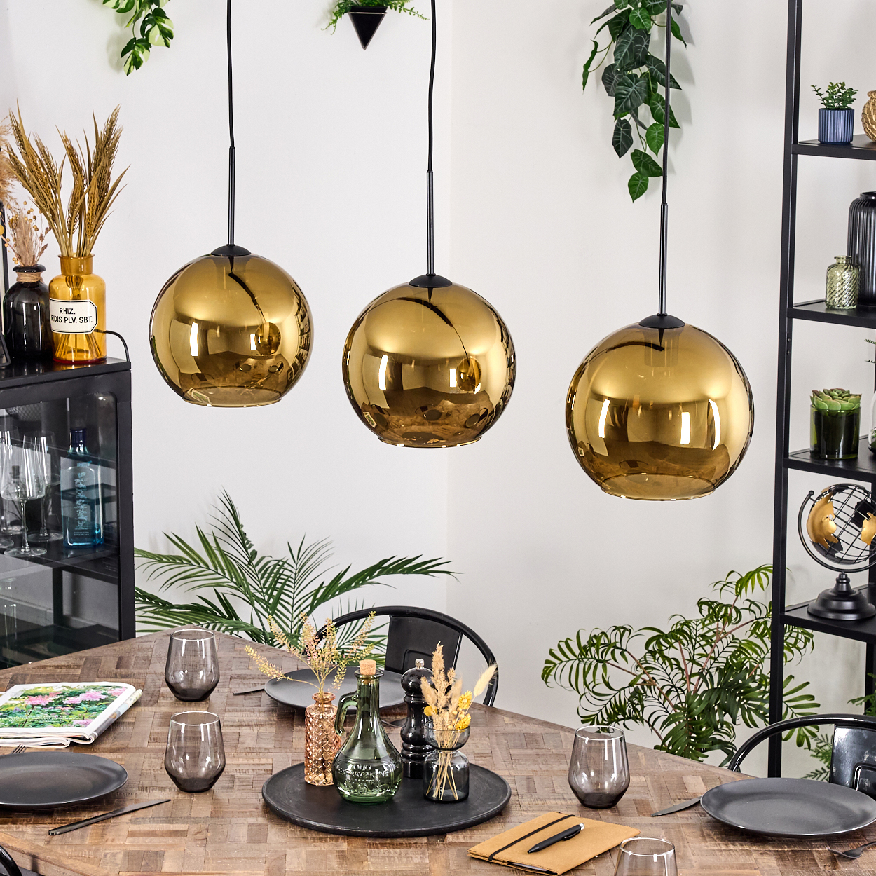 Drei goldene Pendelleuchten über einem Holzesstisch mit Tellern, Gläsern und Dekoration. Pflanzen und Regale im Hintergrund.