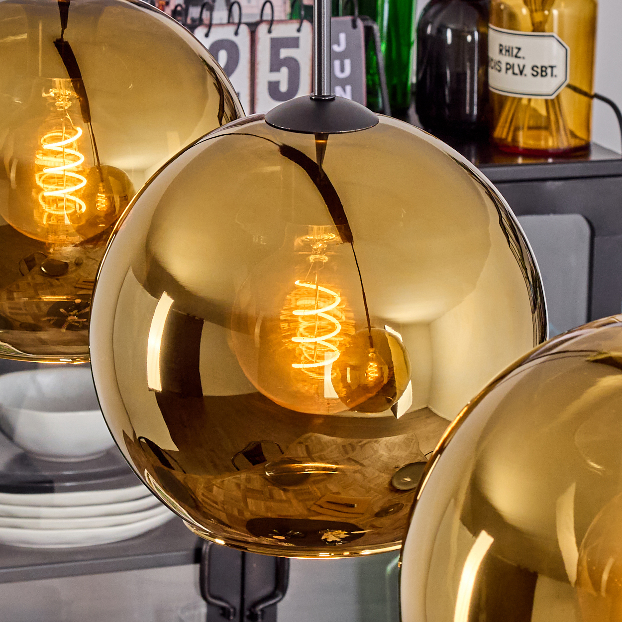 Drei goldene, kugelförmige Pendelleuchten mit leuchtenden Wendelbirnen. Hintergrund: Kalender, Flaschen und weißer Tellerstapel.