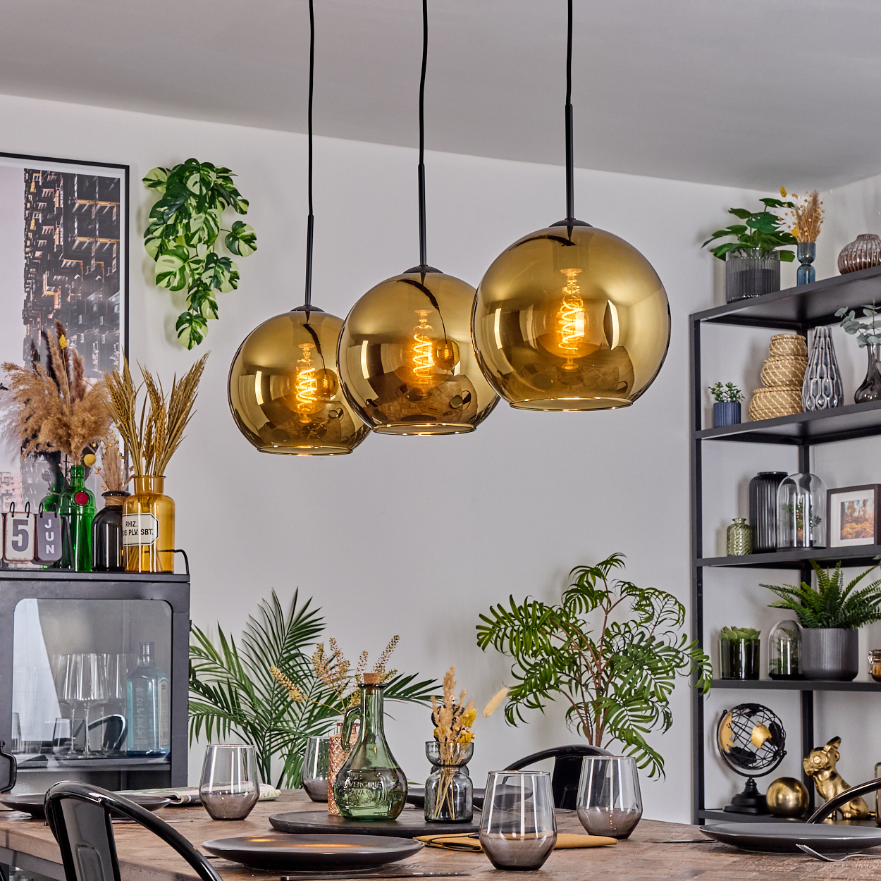 Drei goldene Kugelleuchten über einem Holzesstisch mit Gläsern und Tellern. Pflanzen und Dekoration im Hintergrund.