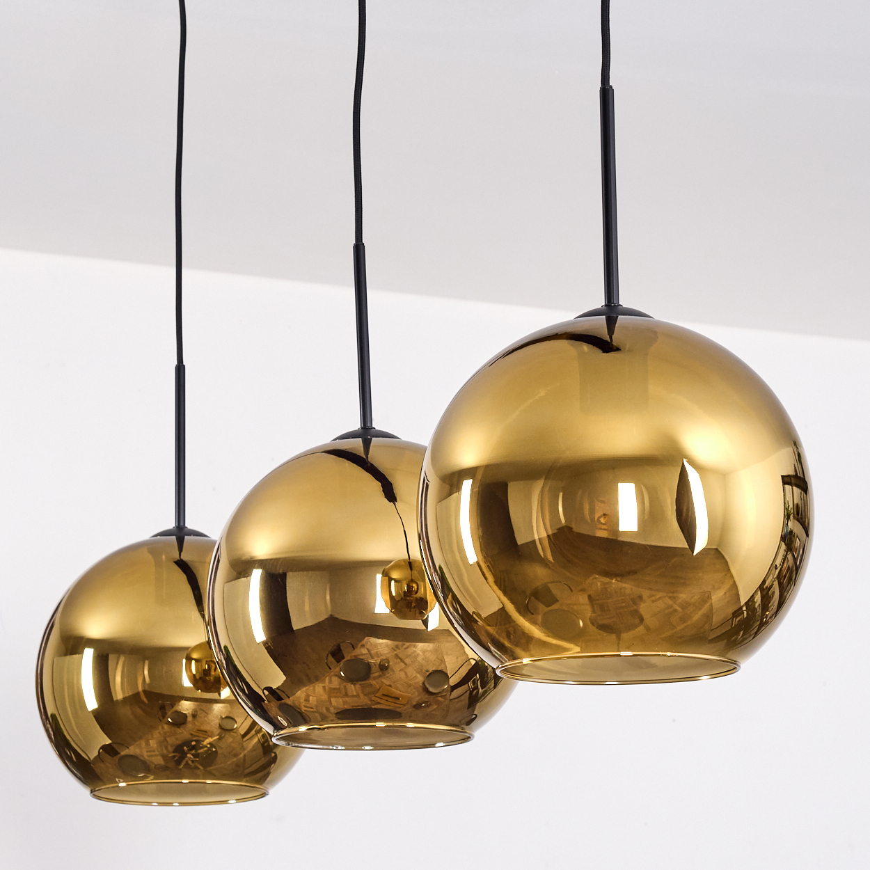 Drei goldene, kugelförmige Pendelleuchten, die an schwarzen Schnüren von einer weißen Decke hängen. Sie reflektieren die Umgebung.