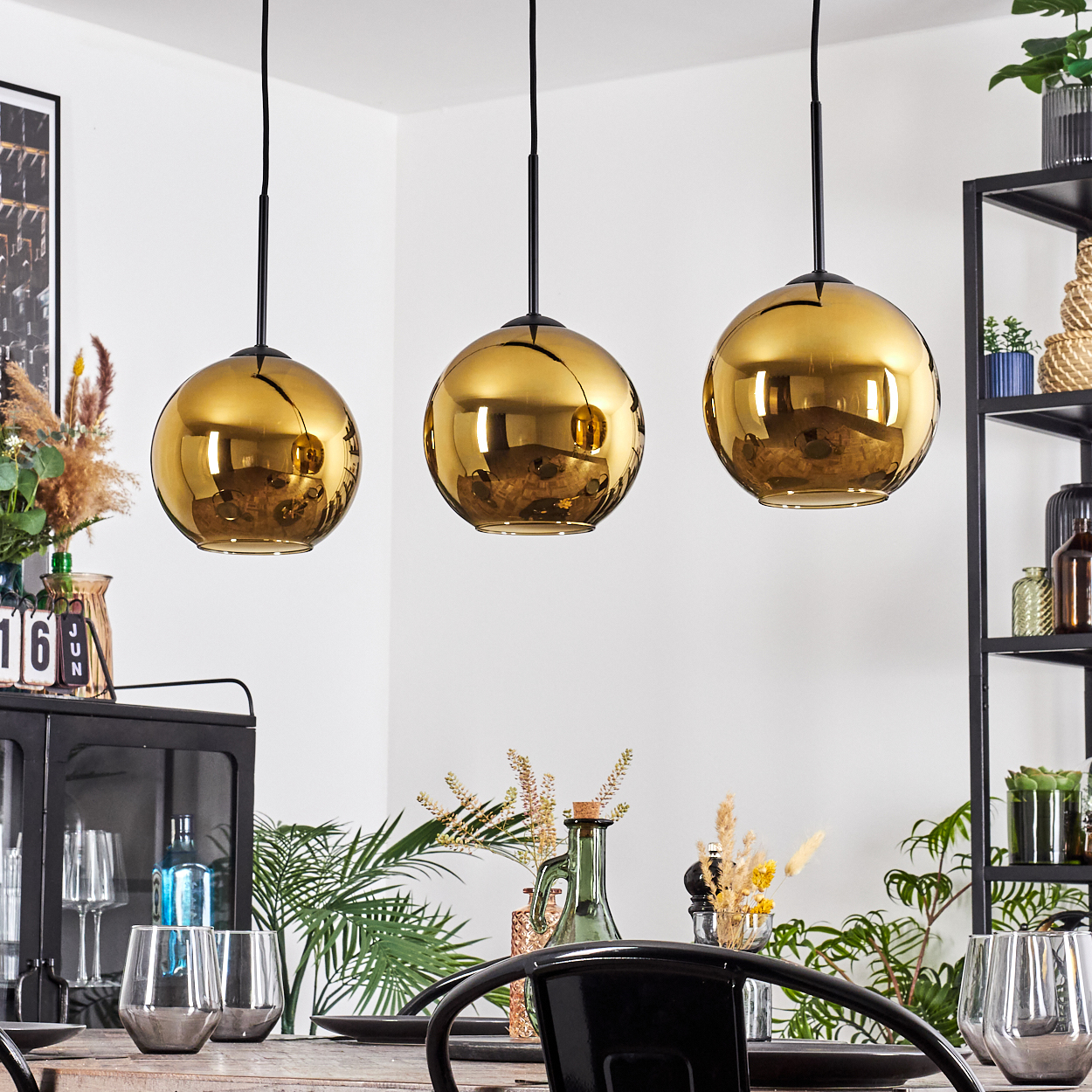 Drei goldene Pendelleuchten hängen über einem Esstisch mit Gläsern und Pflanzen. Rechts ist ein schwarzes Metallregal.
