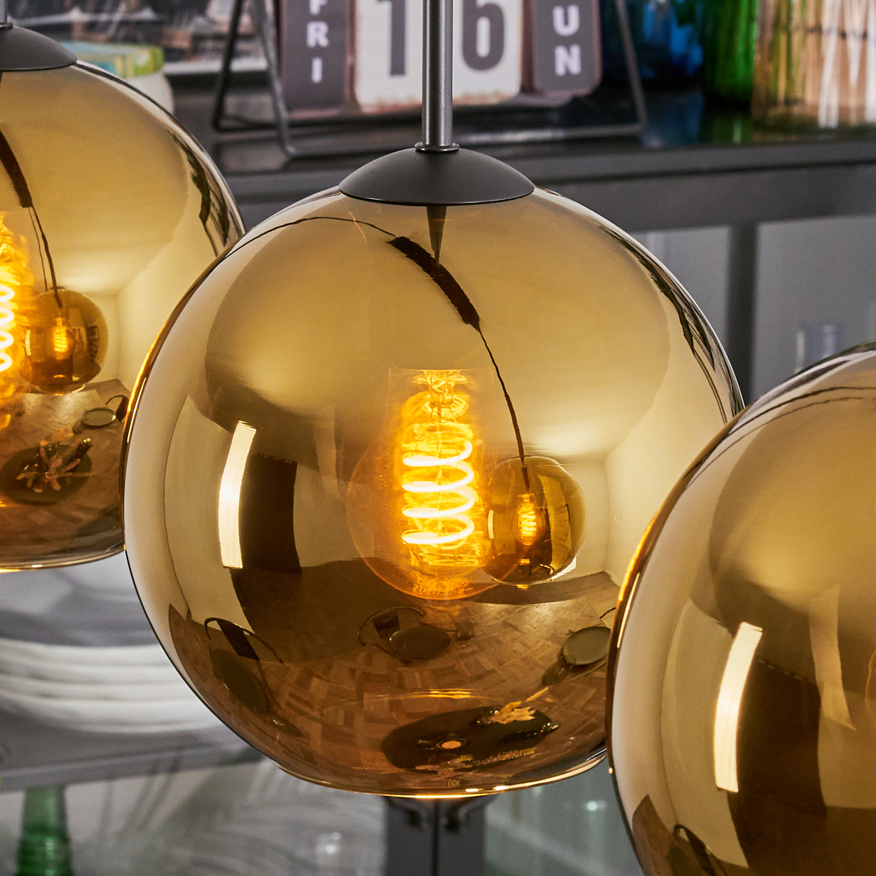 Drei goldene kugelförmige Pendelleuchten mit sichtbaren Filamenten. Im Hintergrund sind ein Kalender und Glasflaschen zu sehen.