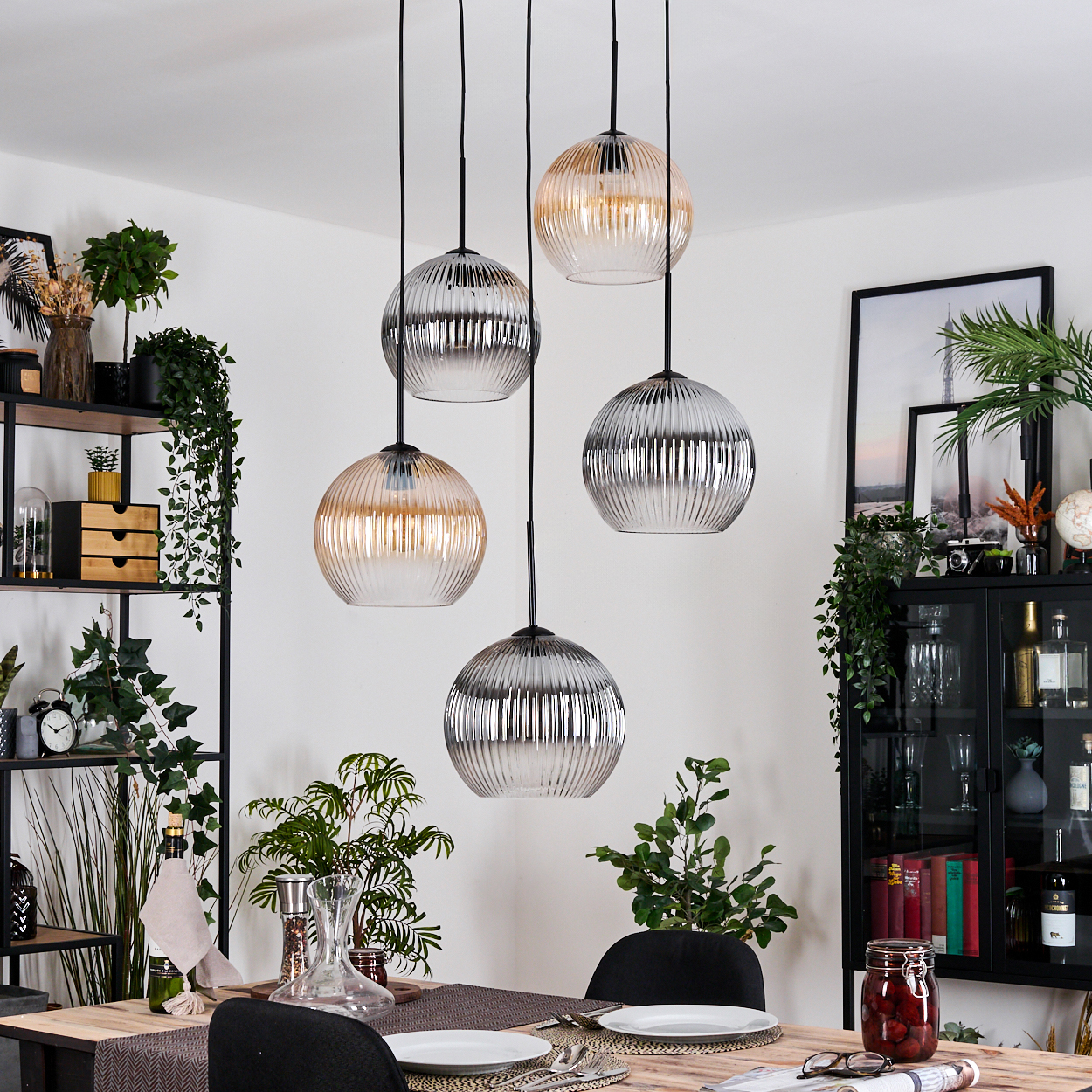 Esszimmer mit Pendelleuchten. Glaslampen, Tisch mit Tellern, Pflanzen und schwarzem Schrank mit Rahmen und Dekor.