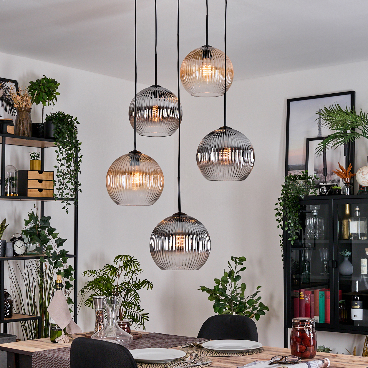 Esszimmer mit gedecktem Tisch. Fünf kugelförmige Pendelleuchten hängen von der Decke. Regale mit Pflanzen und Dekoration sind sichtbar.