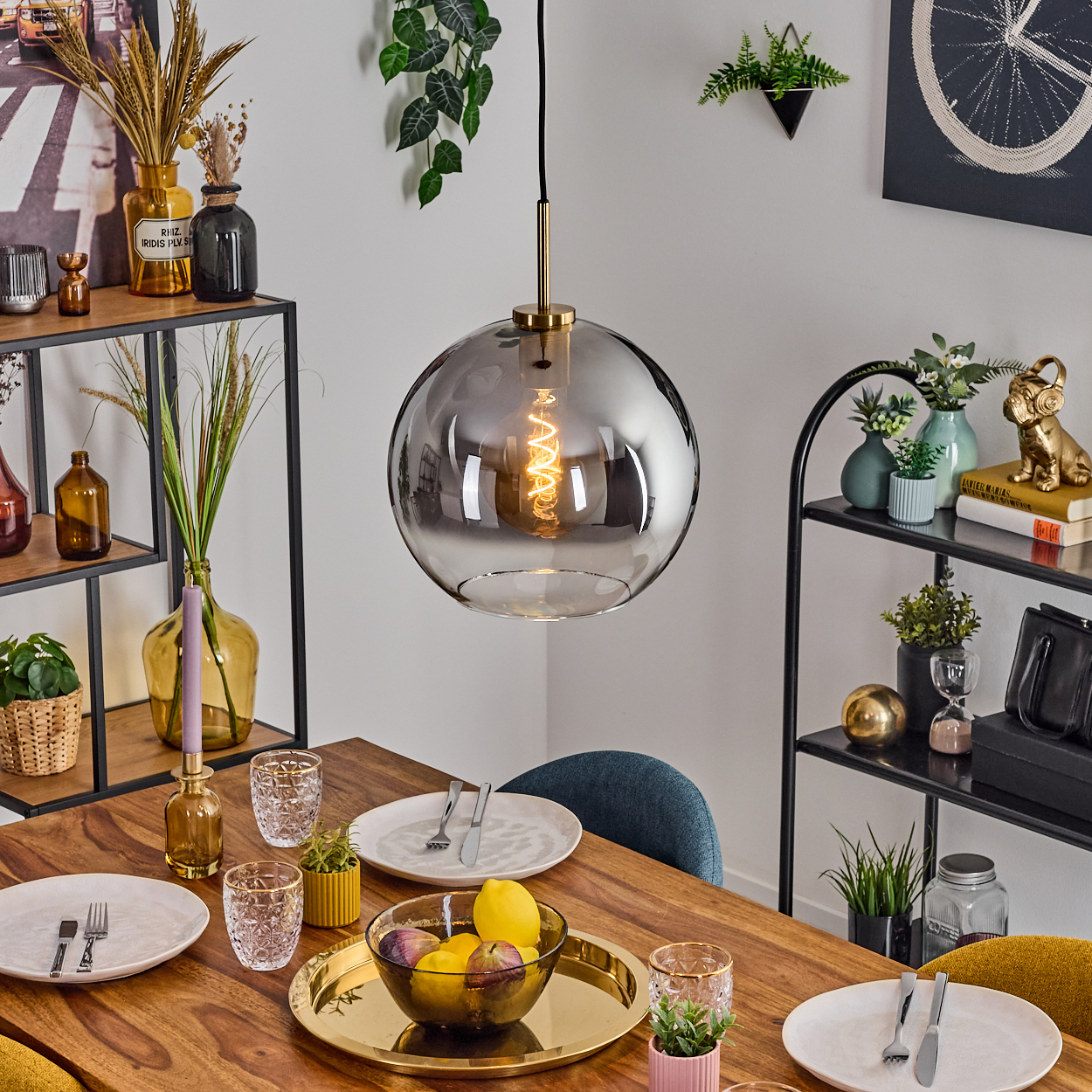 Ein Esstisch ist für eine Mahlzeit gedeckt. Ein großes Glaslicht hängt darüber. Früchte sind in einer Schüssel. Regale mit Dekorationen sind an den Seiten.