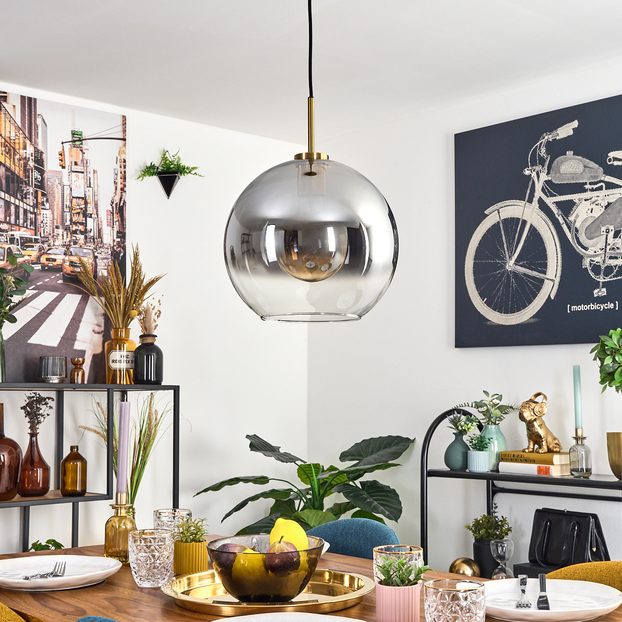 Ein Esszimmer mit einer großen, kugelförmigen Lampe. Ein Tisch ist mit Obst und Gläsern gedeckt. Kunstwerk einer Stadt und eines Fahrrades.