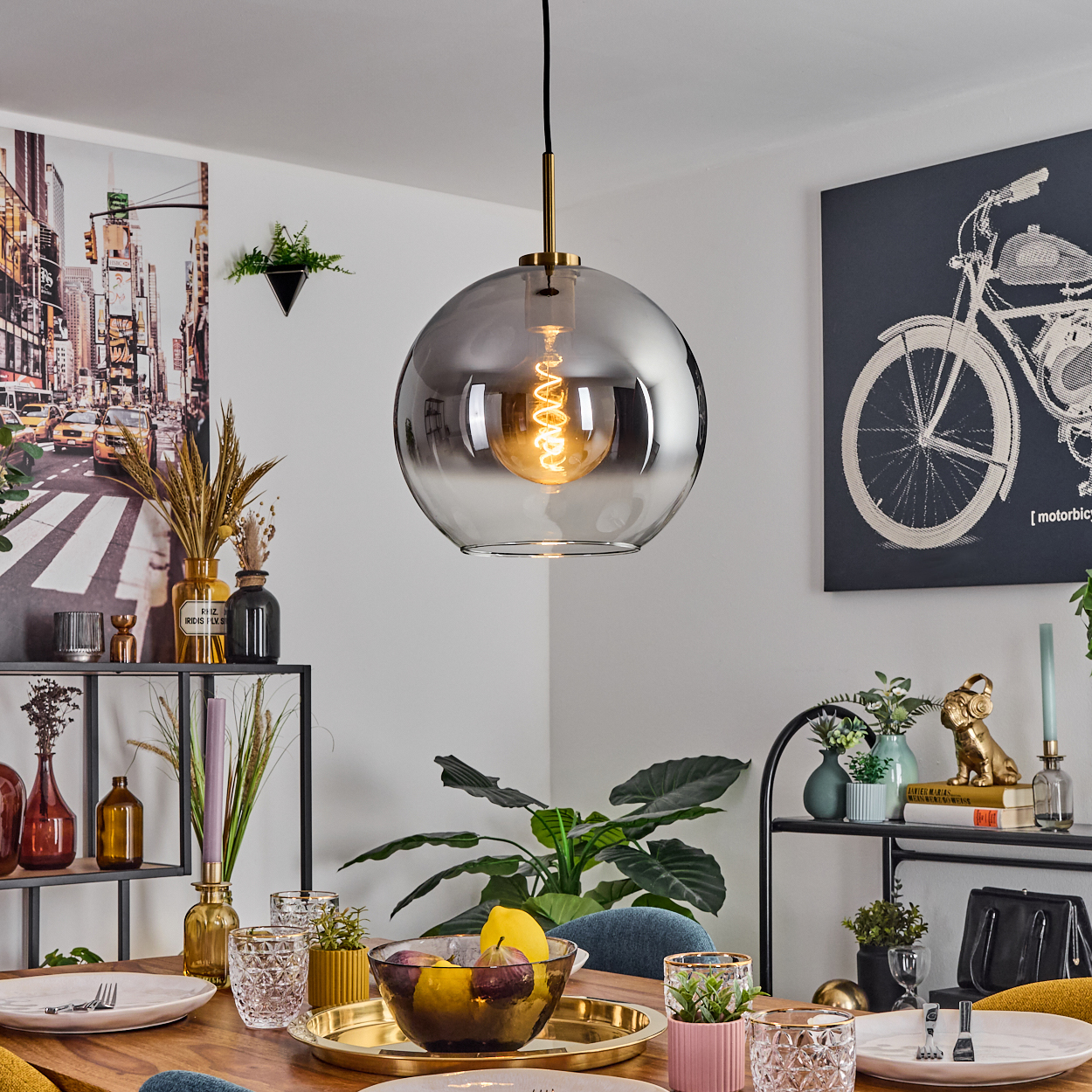 Ein Esszimmer mit Hängelampe. Ein Tisch ist mit Tellern, Gläsern und einer Obstschale gedeckt. Kunstwerke und Pflanzen schmücken die Wände.