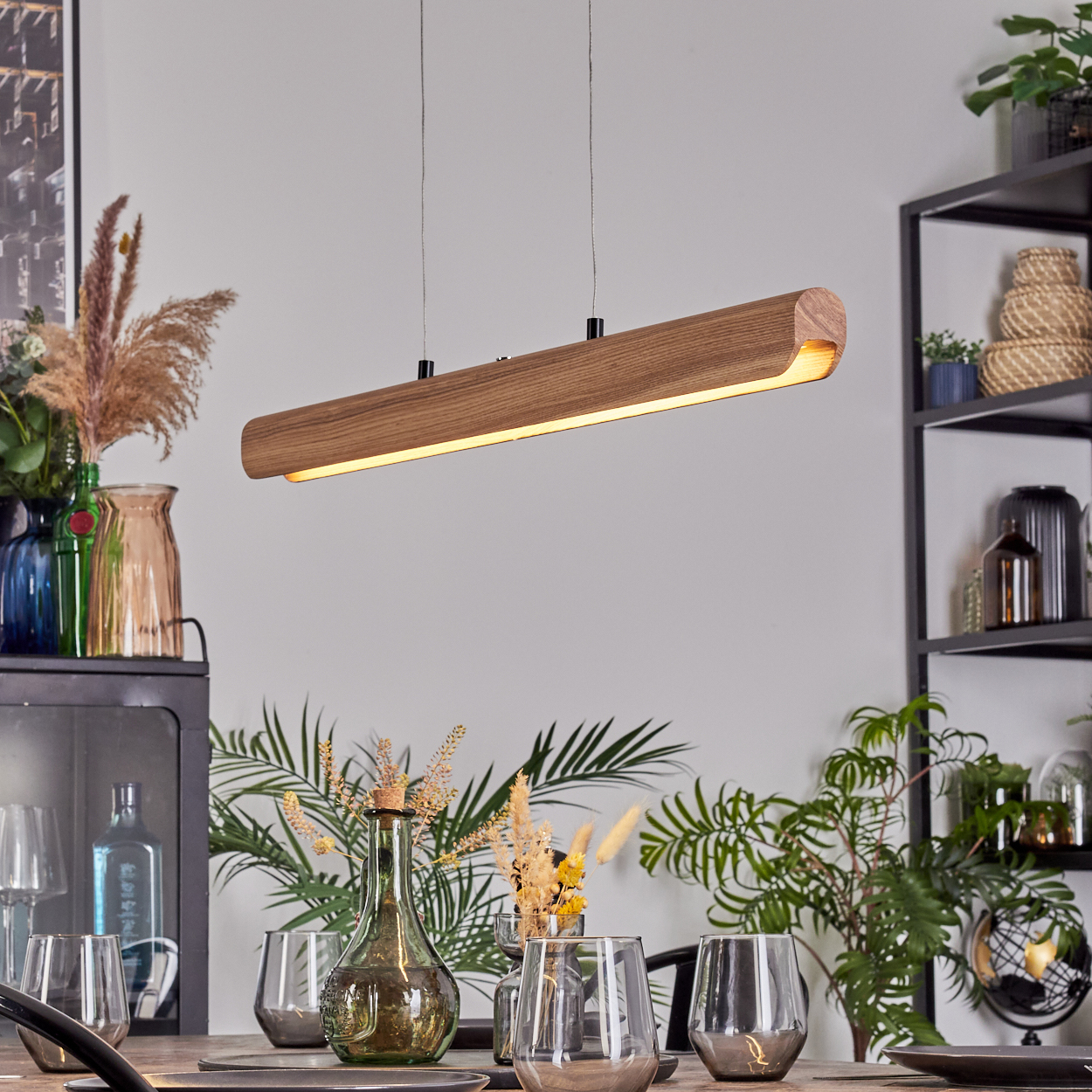 Eine Holzlampe hängt über einem Tisch mit Gläsern und Tellern. Pflanzen und Dekorationsartikel befinden sich im Hintergrund.