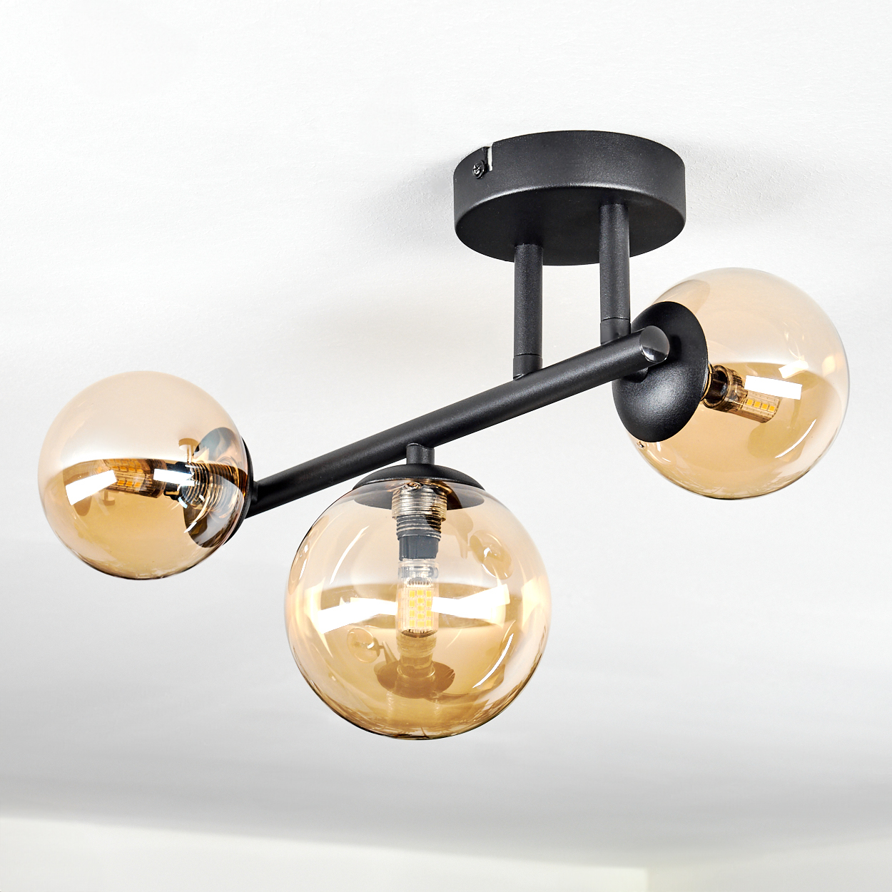 Eine moderne Deckenleuchte mit drei bernsteinfarbenen Glaskugeln an einem schwarzen Metallrahmen, montiert an einer weißen Decke. Jede Glühbirne leuchtet.