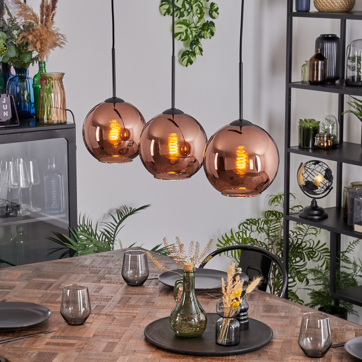 Drei kupferfarbene Pendelleuchten hängen über einem Holztisch, der für das Abendessen gedeckt ist. Vasen und Dekorationen befinden sich im Hintergrund.