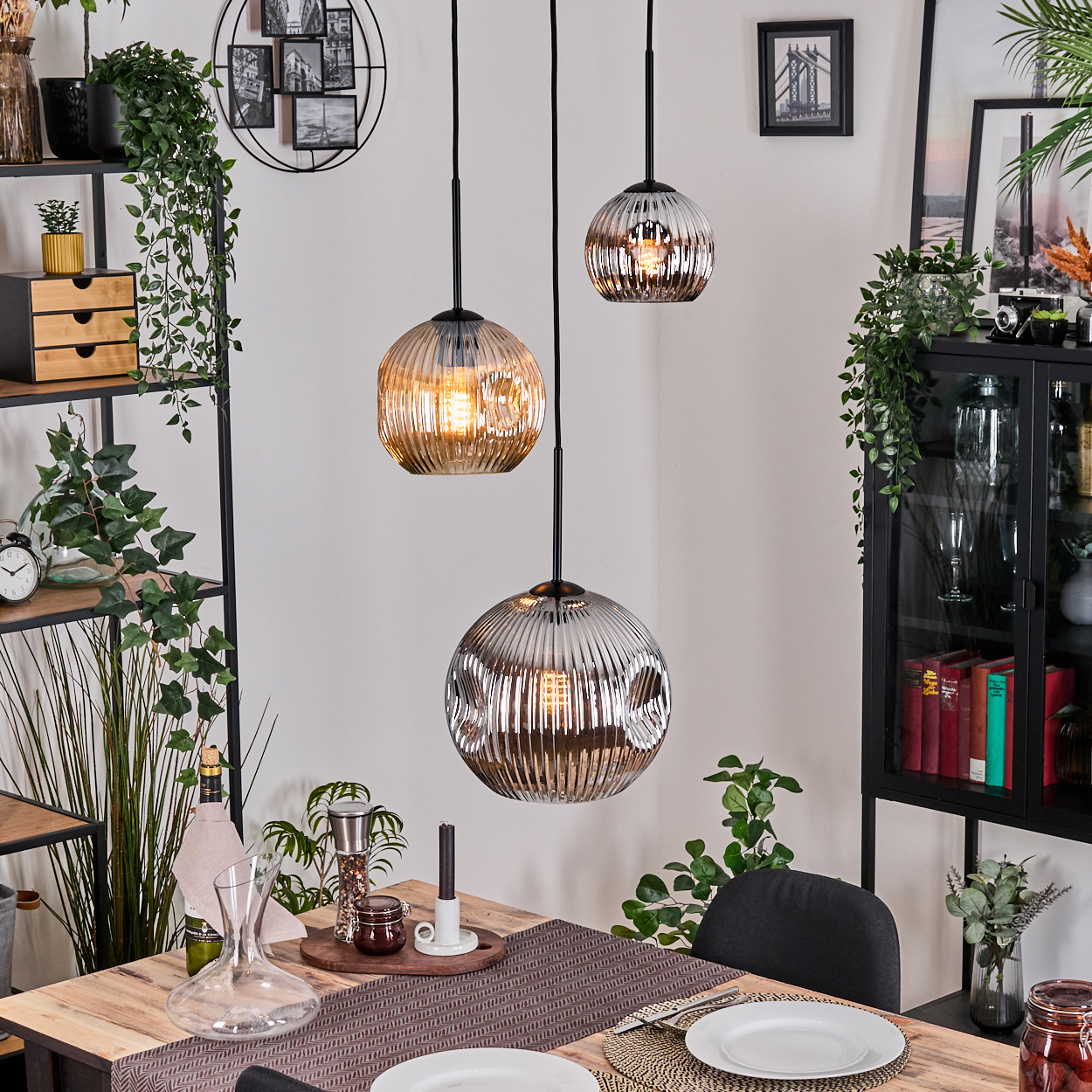 Drei Pendelleuchten hängen über einem Holztisch, der für eine Mahlzeit gedeckt ist. Im Hintergrund Regale mit Pflanzen und Dekoration.