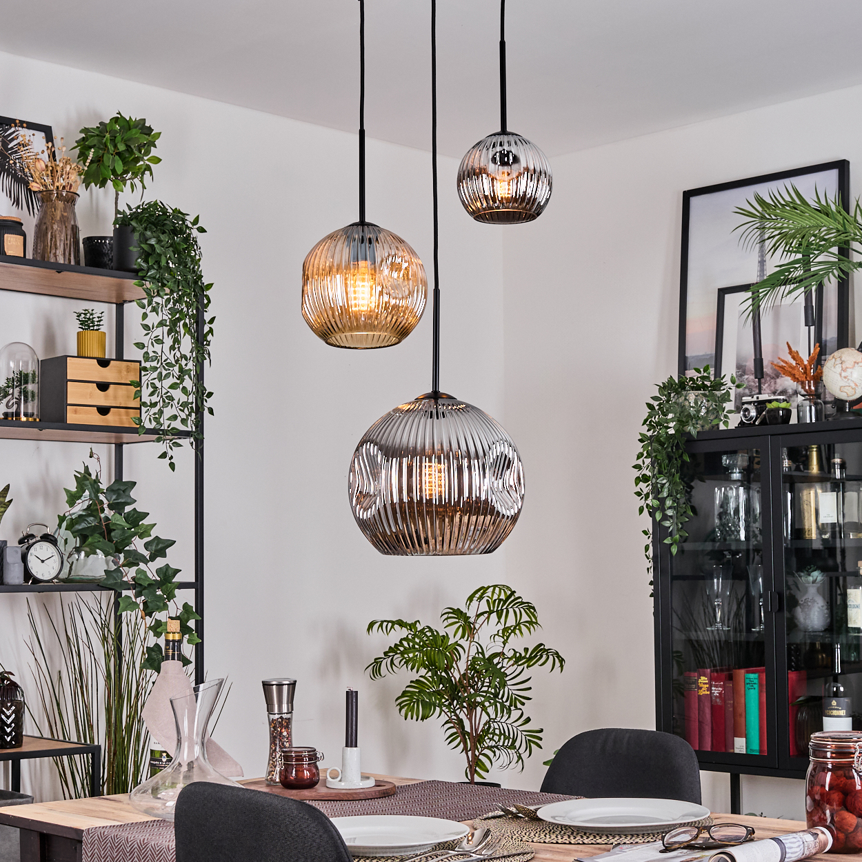 Esszimmer mit drei Hängelampen. Tisch mit Tellern und Gläsern gedeckt. Regale und Schrank mit Pflanzen und Dekorationsartikeln.