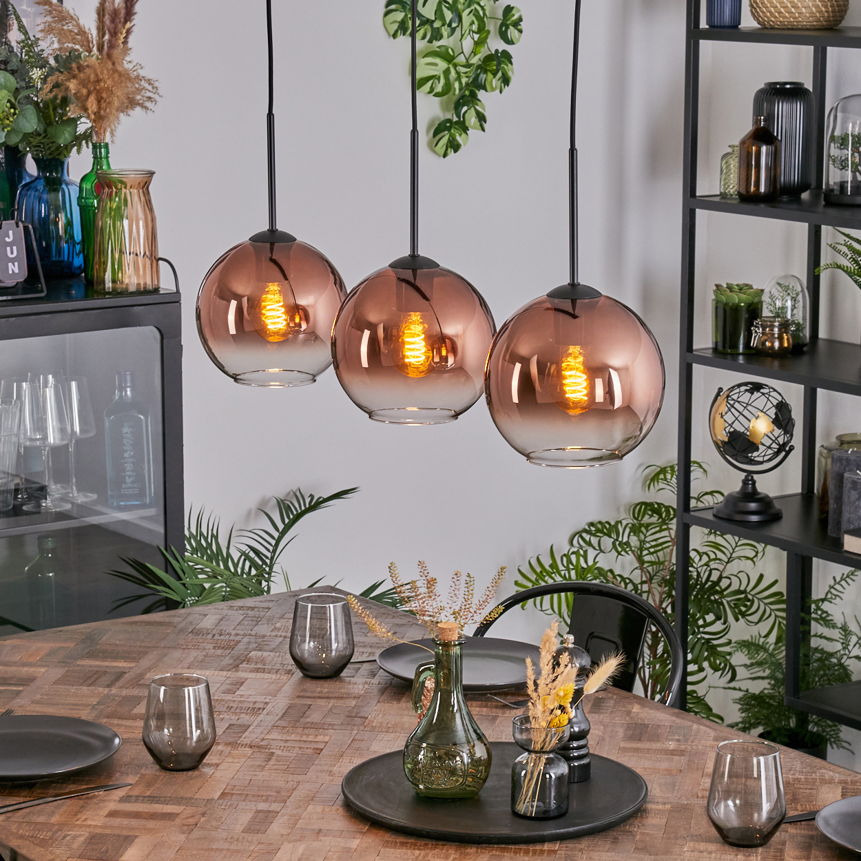 Esszimmer mit drei Hängelampen. Holztisch mit Gläsern und Dekor. Regale mit Pflanzen und Dekor im Hintergrund.