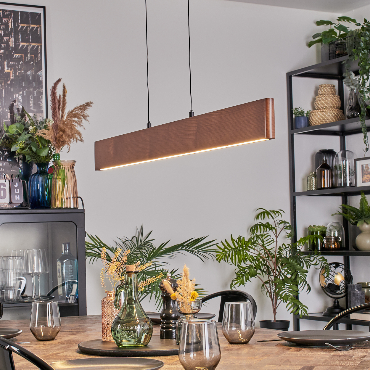 Esszimmer mit Holztisch, Gläsern, dekorativen Pflanzen und einer langen Holzlampe. Regale mit Pflanzen im Hintergrund.