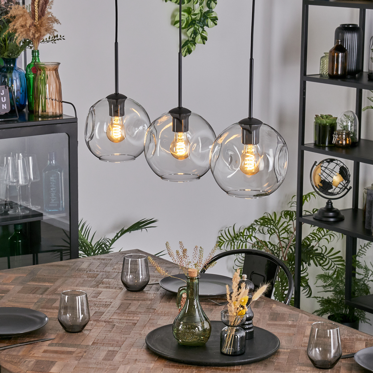 Esszimmer mit Holztisch, drei Hängelampen und Dekorationsartikeln. Inklusive Gläsern, Pflanzen und einem schwarzen Regal.