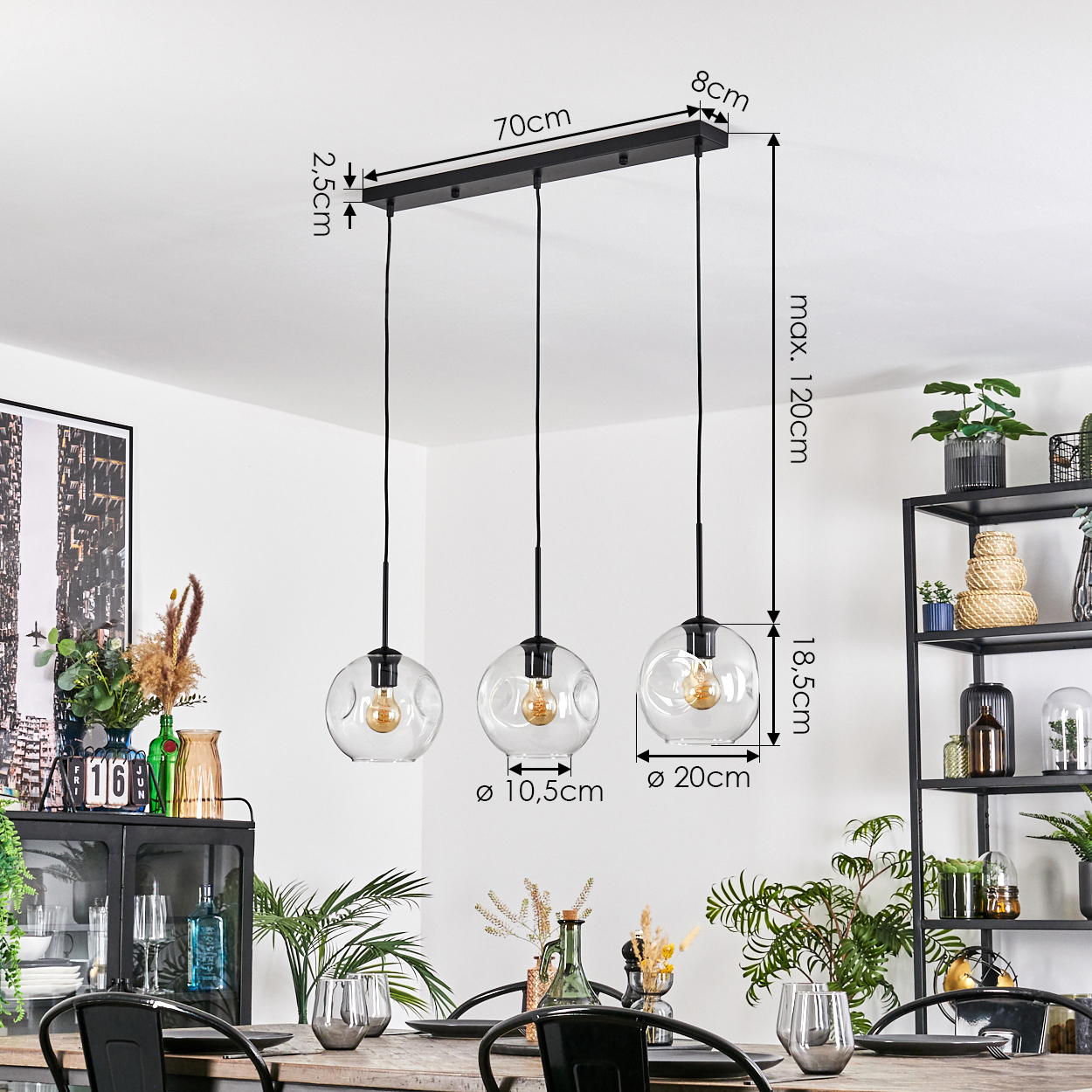 Drei Pendelleuchten mit Glaskugeln hängen über einem Holztisch. Abmessungen sind beschriftet. Pflanzen und ein schwarzes Metallregal im Hintergrund.
