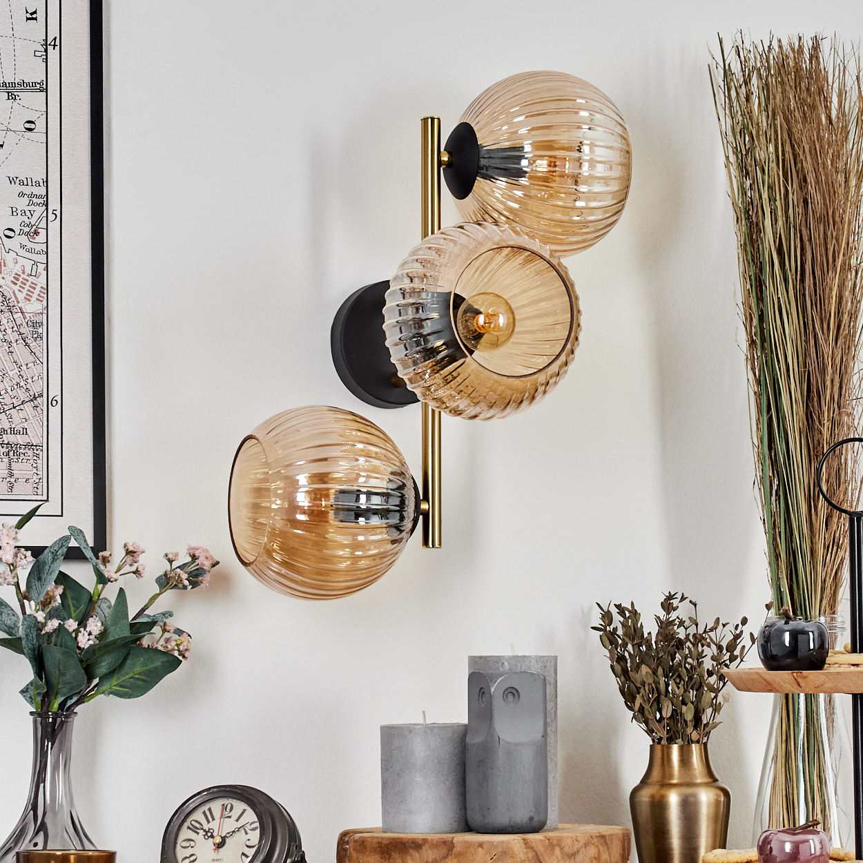 Eine Wandlampe mit drei bernsteinfarbenen Glaskugeln, einem schwarzen Sockel und goldenen Akzenten. Daneben befinden sich eine Karte, Vasen, Kerzen und Dekoration.