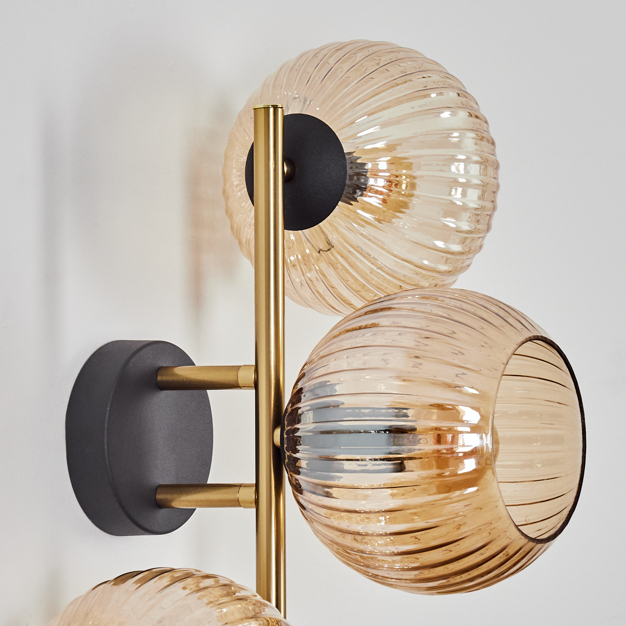Eine moderne Wandlampe mit drei bernsteinfarbenen Glaskugelschirmen. Die Lampe hat ein goldfarbenes Gestell und einen schwarzen Sockel.