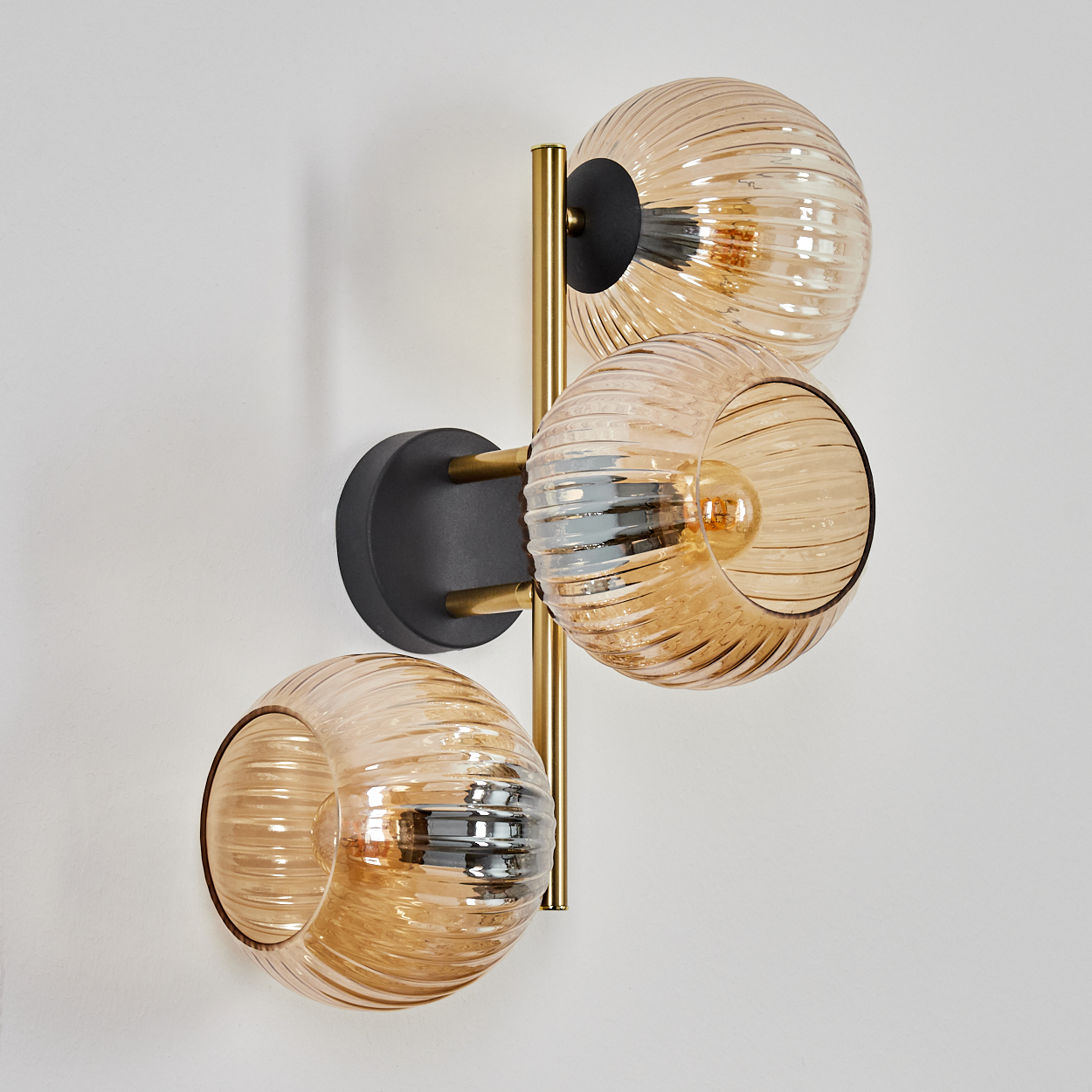 Eine Wandlampe mit drei bernsteinfarbenen Glaskugeln. Die Lampe hat einen goldfarbenen Rahmen und einen schwarzen Sockel, an einer weißen Wand montiert.