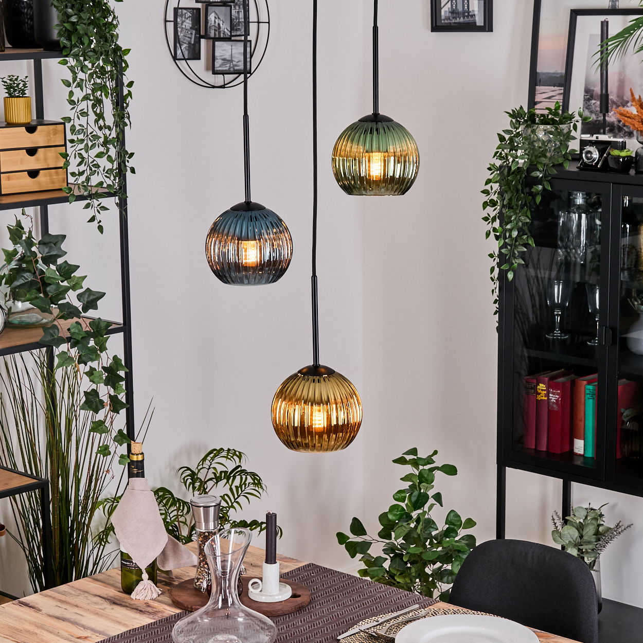 Drei Pendelleuchten über einem Holztisch. Pflanzen und Dekorationen füllen den Hintergrund. Eine Lampe ist blau, die anderen sind grün und gold.